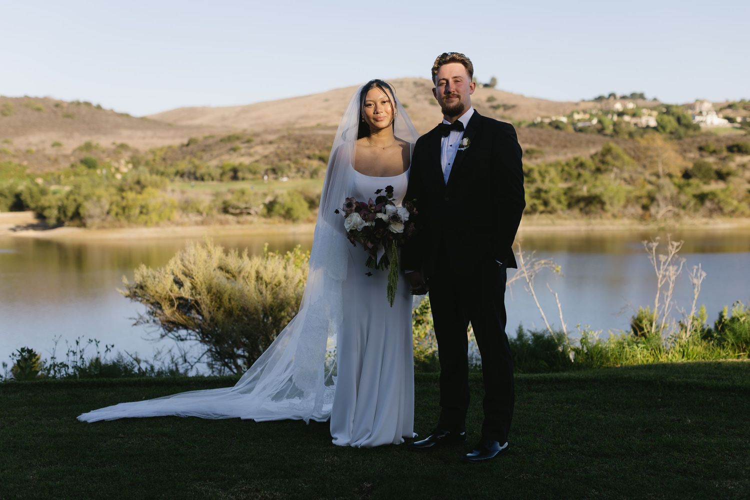 A couple poses for their wedding portrait on a lawn overlooking a serene lake and rolling hills.