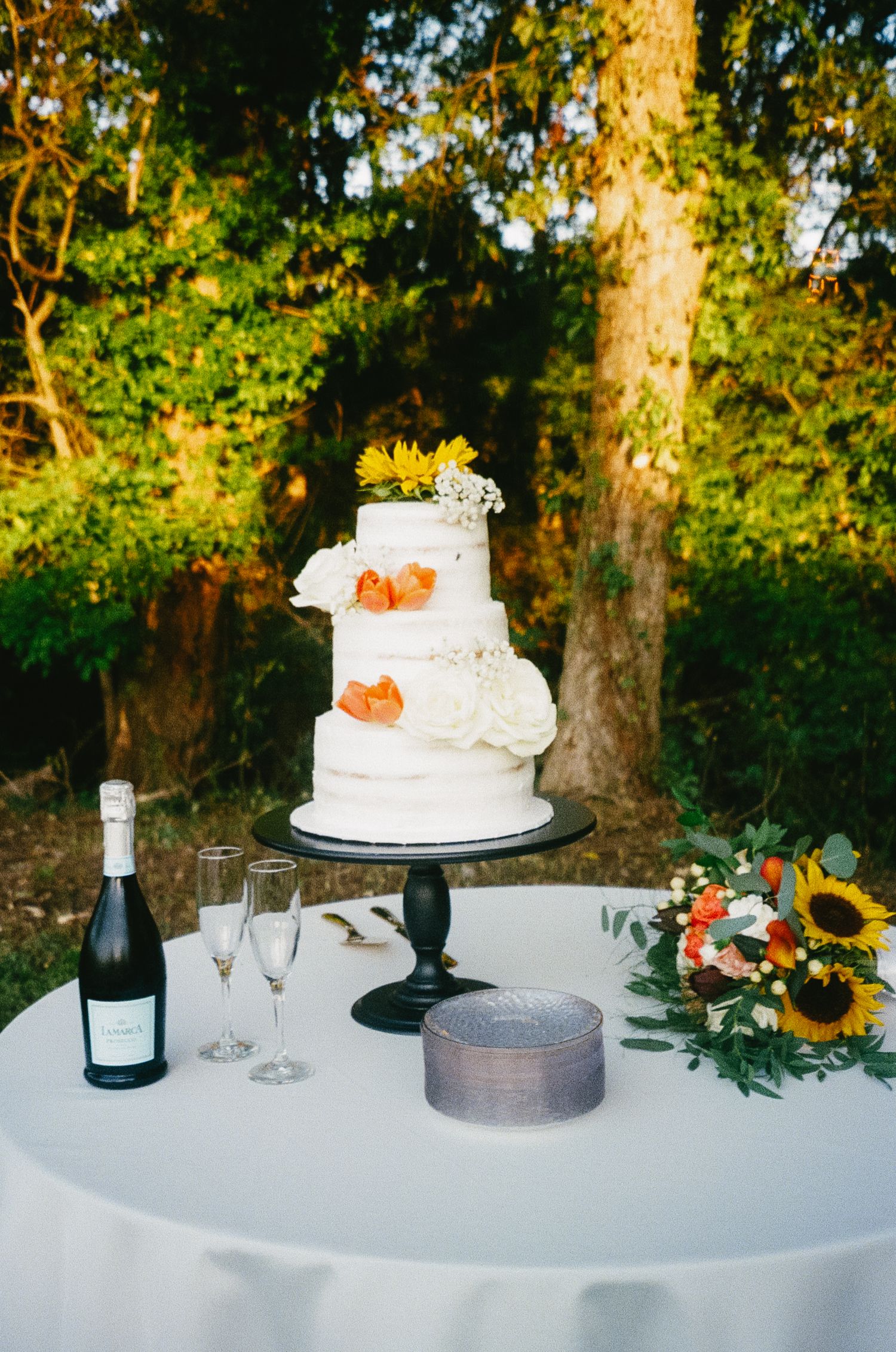 Wedding cake display on white tablecloth with champagne bottle, sunflowers and autumn decor at outdoor reception.