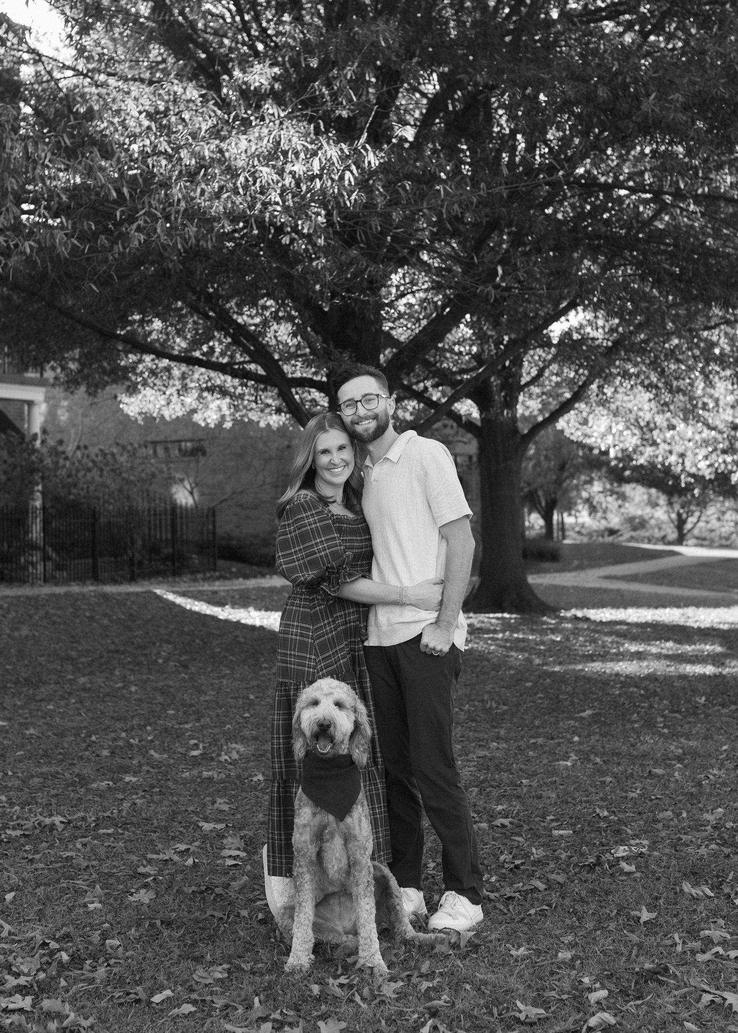 A black and white photo of a couple with their goldendoodle dog under a large tree.