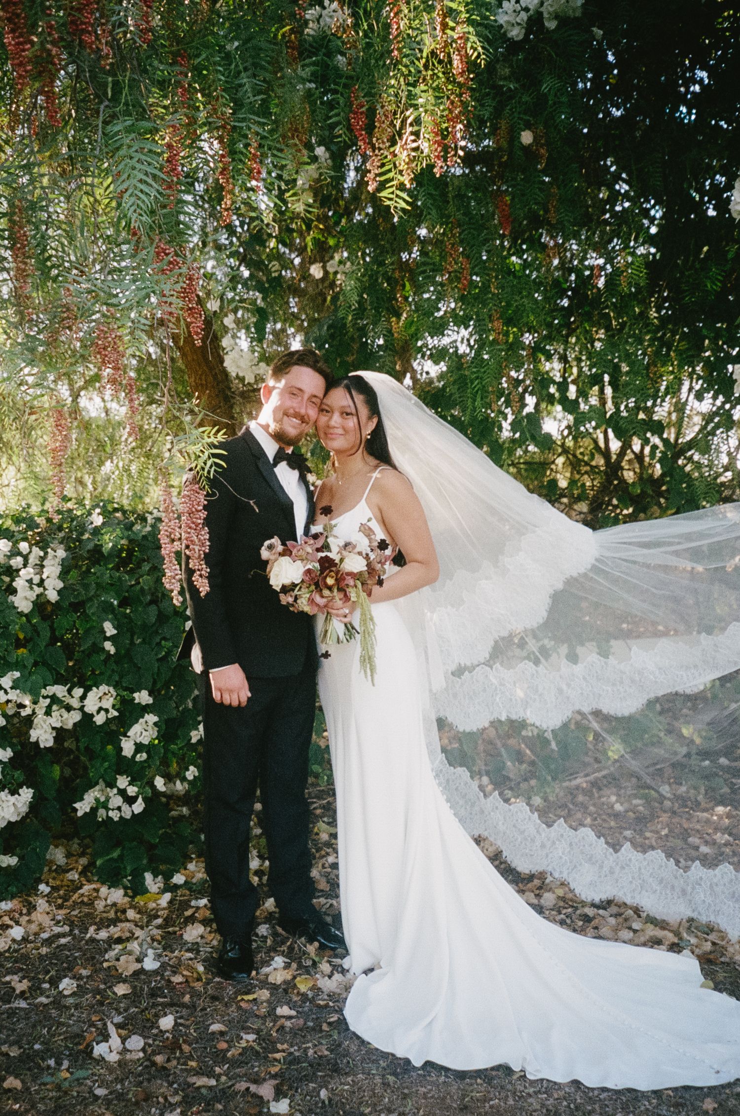 An elegant wedding portrait under flowering trees with cascading blooms and a flowing white veil.