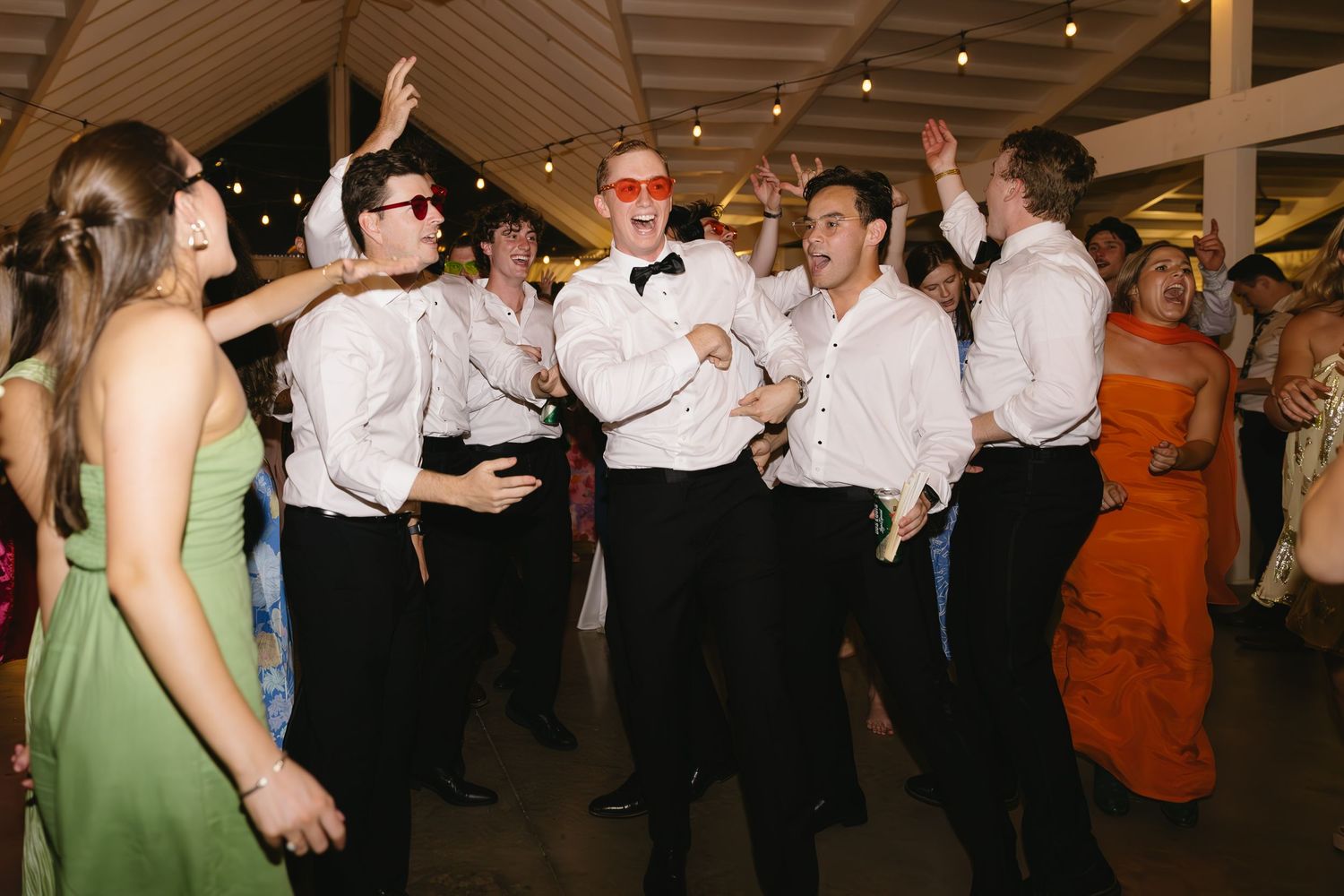 Wedding reception dance floor filled with guests in formal attire dancing under string lights.