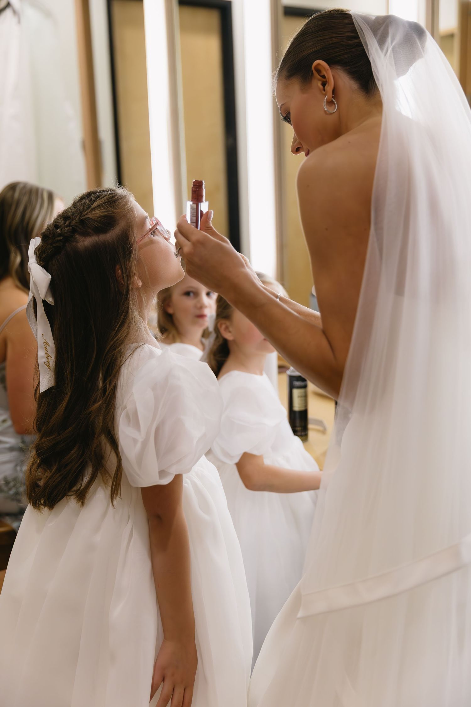 A bride applying makeup to a young attendant in a white dress during wedding preparations.