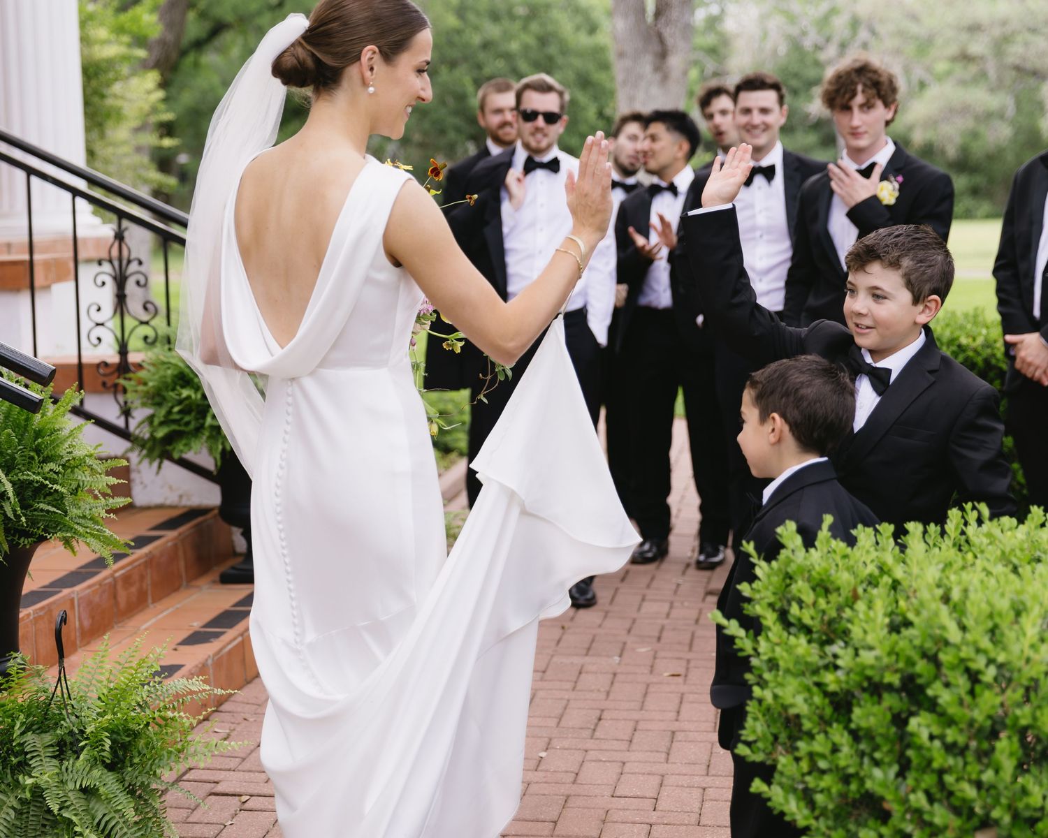 Someone in an elegant white wedding gown stands on brick steps surrounded by formally dressed guests at an outdoor wedding.