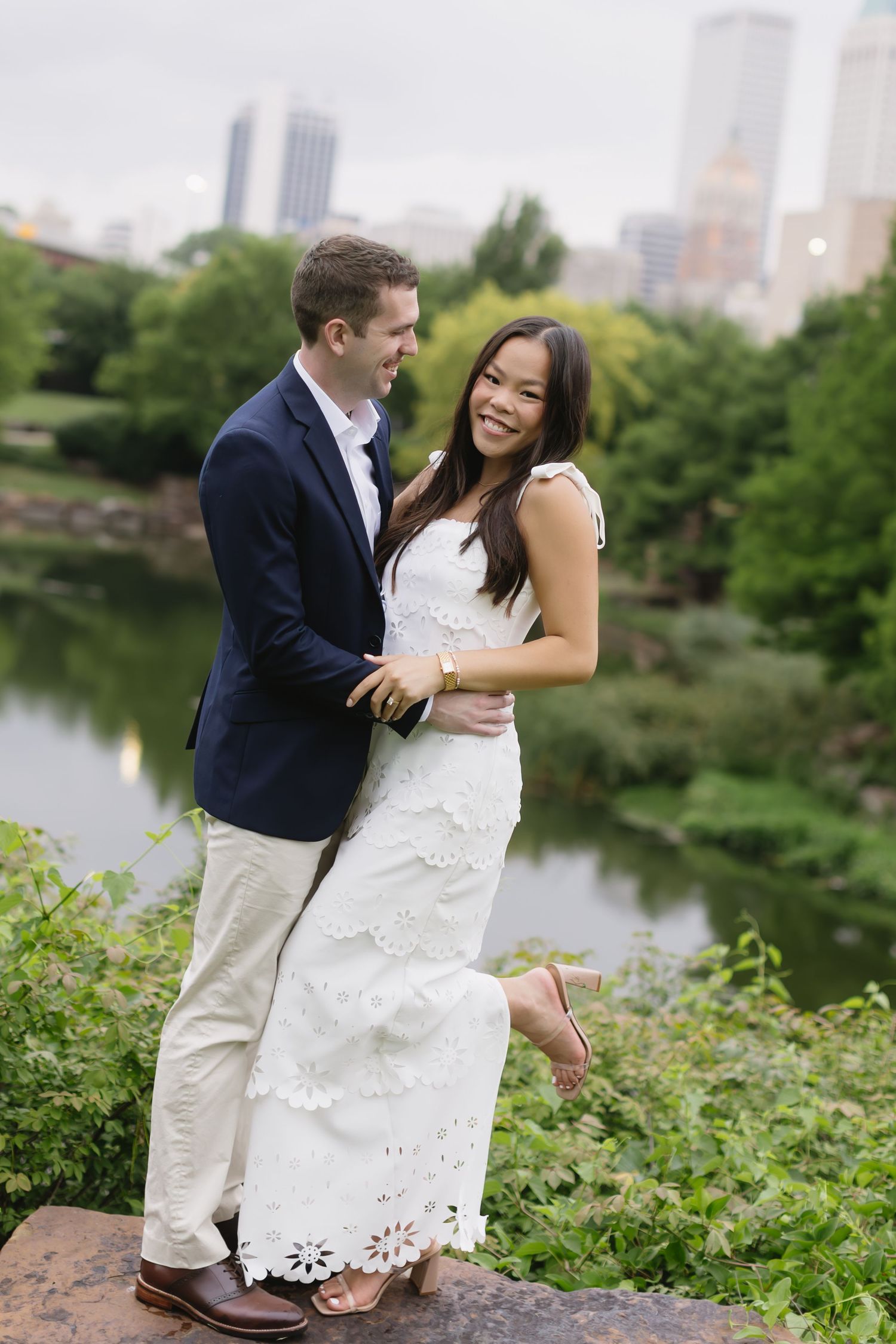 A sweet embrace by the waterfront with greenery and urban buildings creating a romantic backdrop.