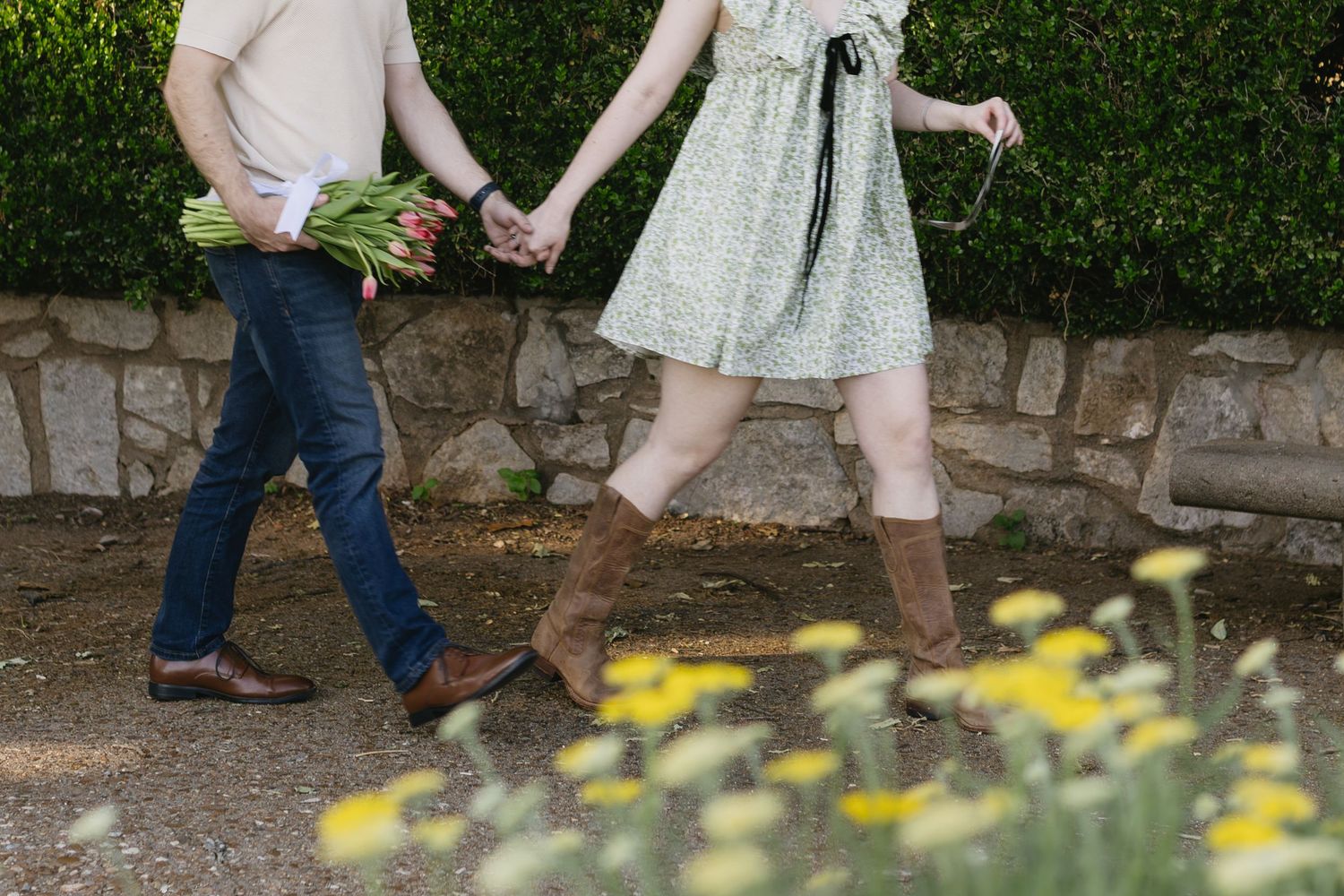 Two people holding hands and dancing together on a stone path surrounded by greenery and yellow flowers.
