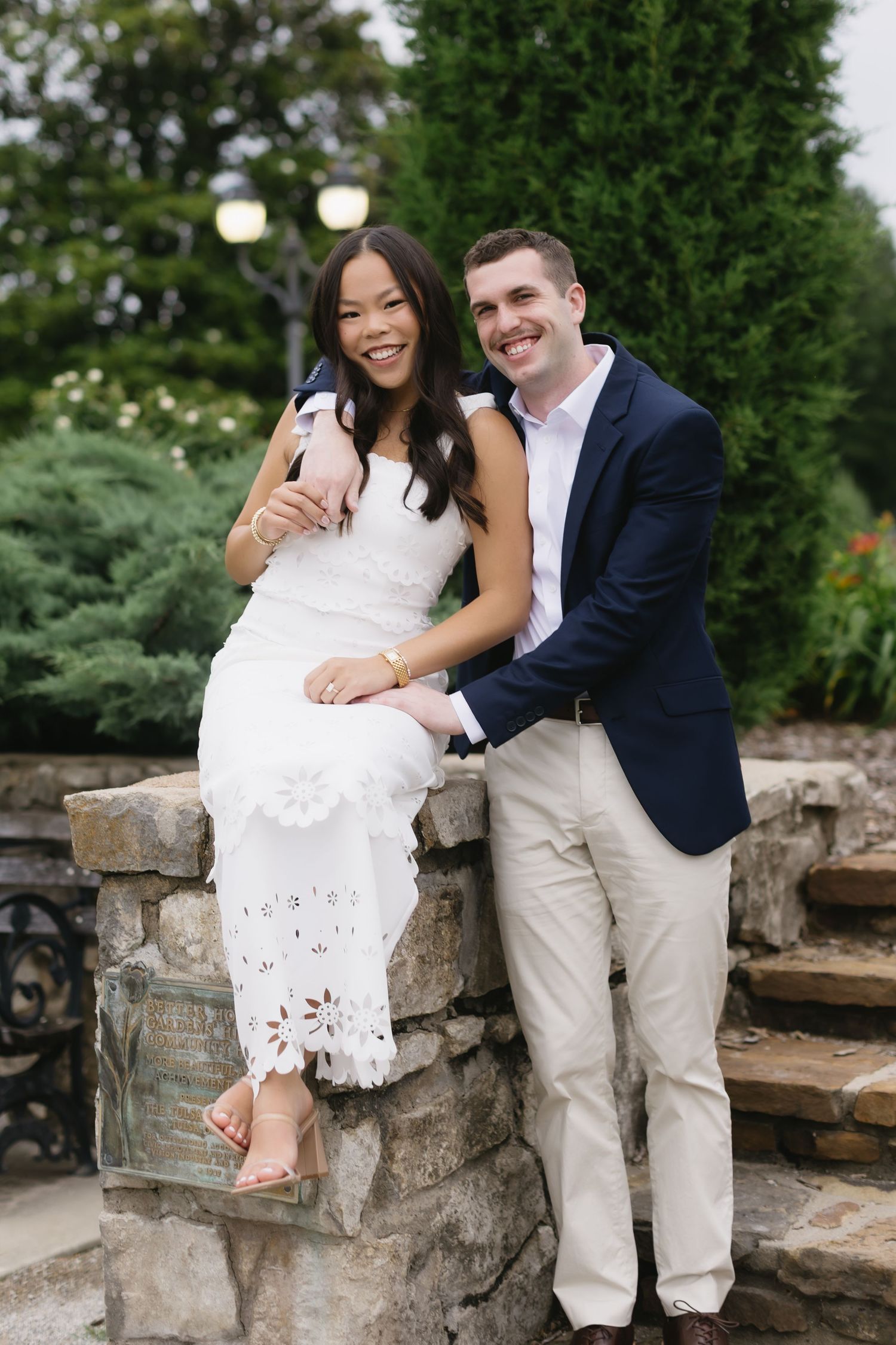 A romantic photoshoot on a stone wall with a couple wearing a white lace dress and navy blazer with khaki pants.