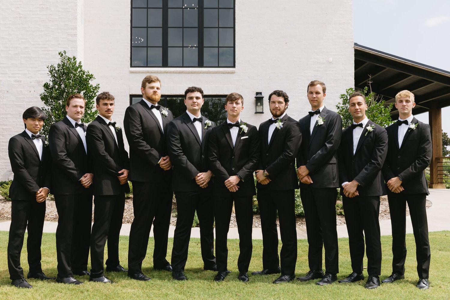 Wide shot of groomsmen lined up in black tuxedos outside a modern church.