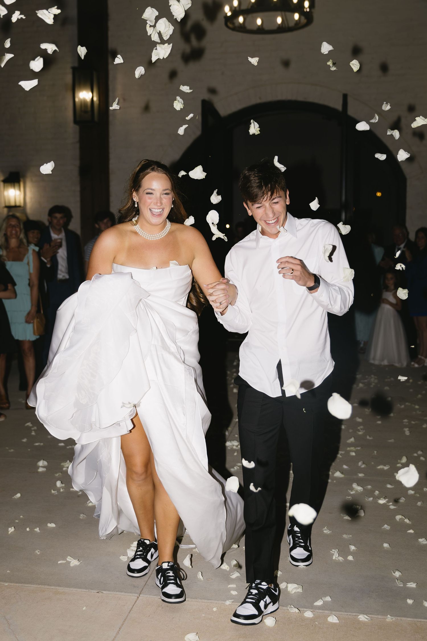 Wedding exit with white flower petals falling as couple runs through in formal attire and sneakers.
