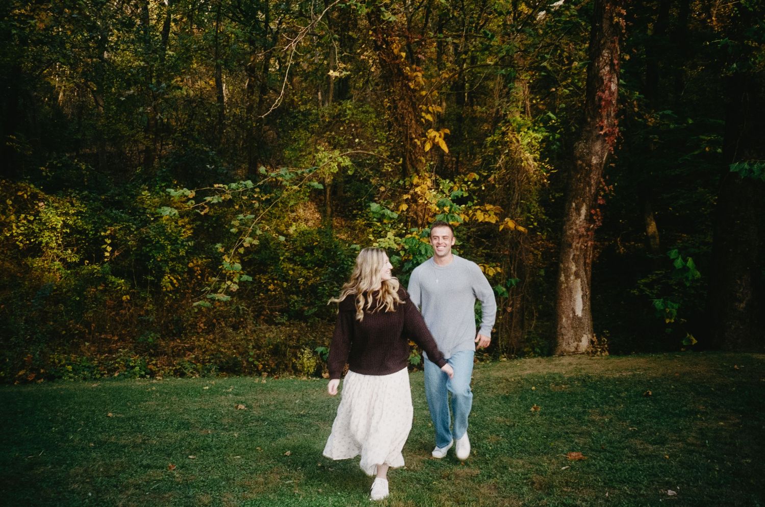 A couple walks hand in hand through a sunlit autumn forest path with golden leaves and tall trees.