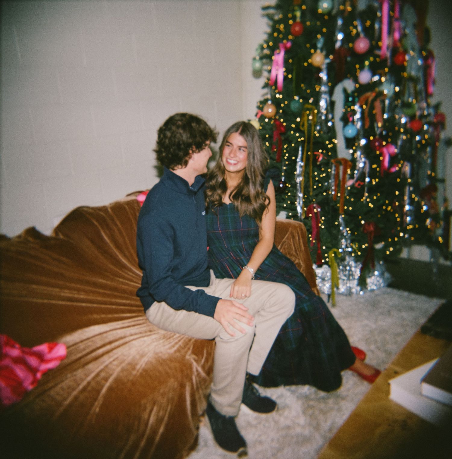 A couple sits together on a brown couch by a decorated Christmas tree with lights and ornaments in a cozy living room.