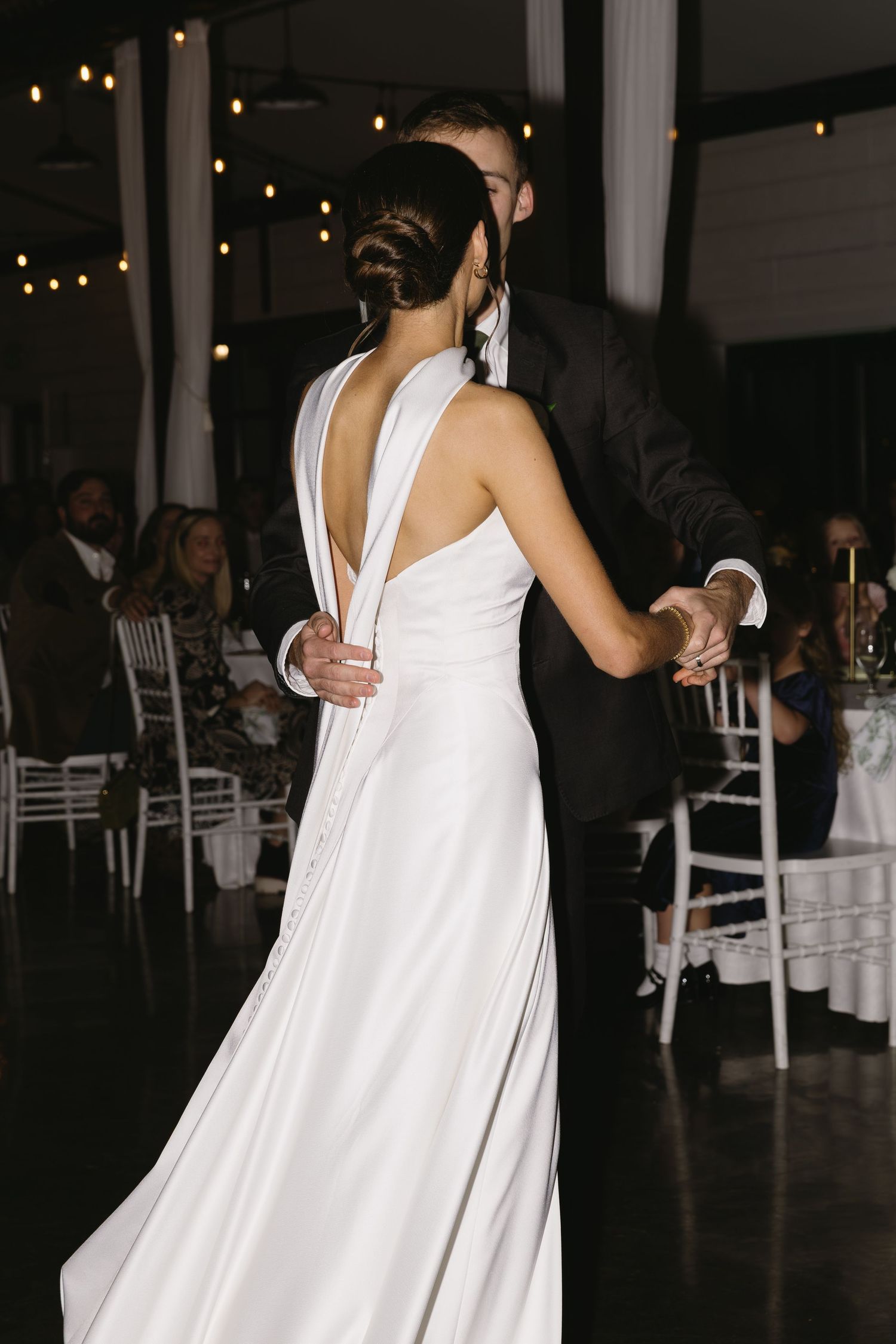 A bride in a flowing white dress dances at a dimly lit wedding reception with string lights.