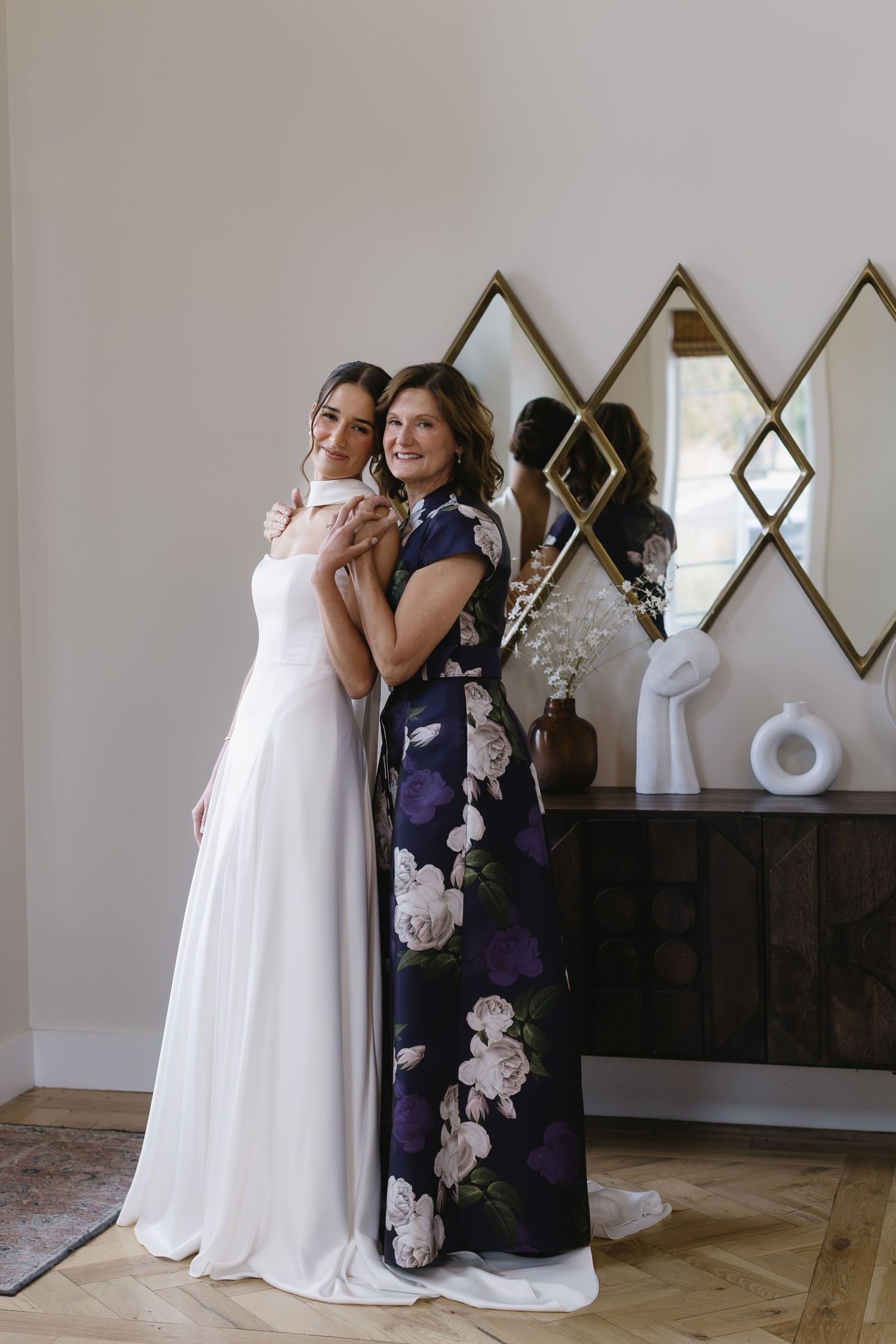 Two people embracing in formal wear against a white wall with diamond-shaped mirrors.
