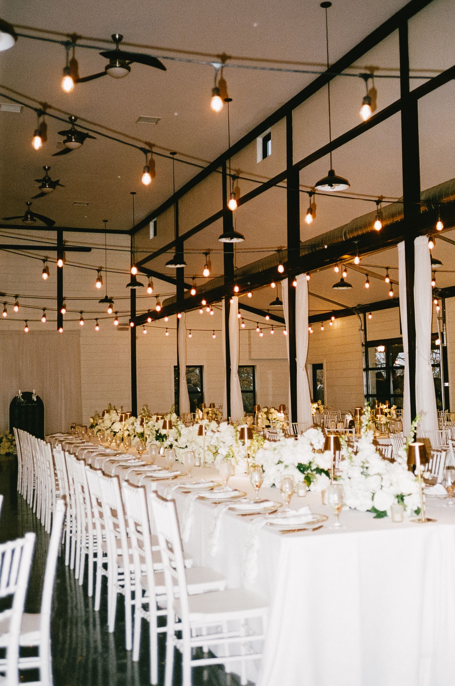 Elegant wedding reception venue with long tables, white chairs, and string lights overhead.