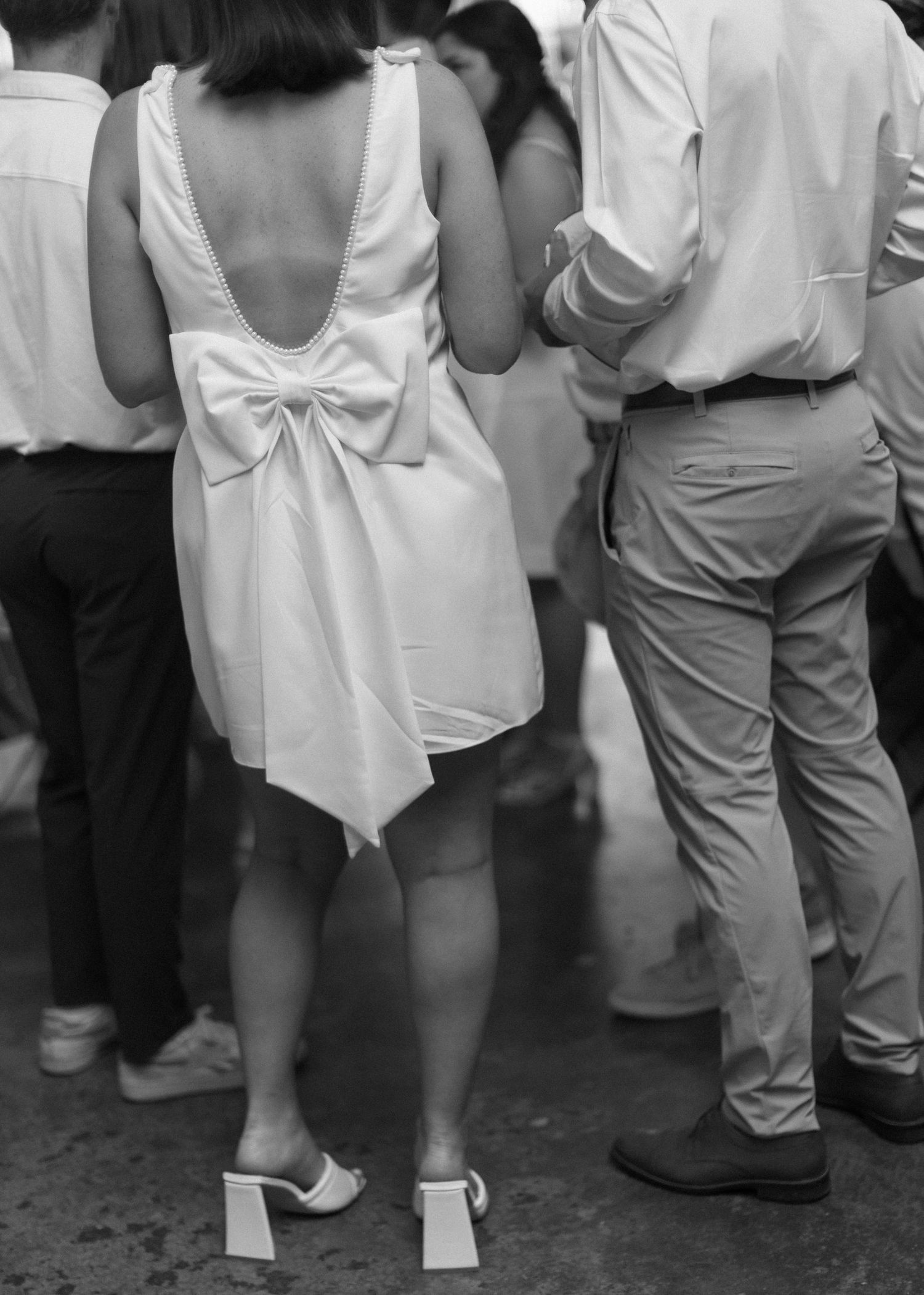 Black and white photo of a woman in white dress with large bow detail on back standing among crowd.