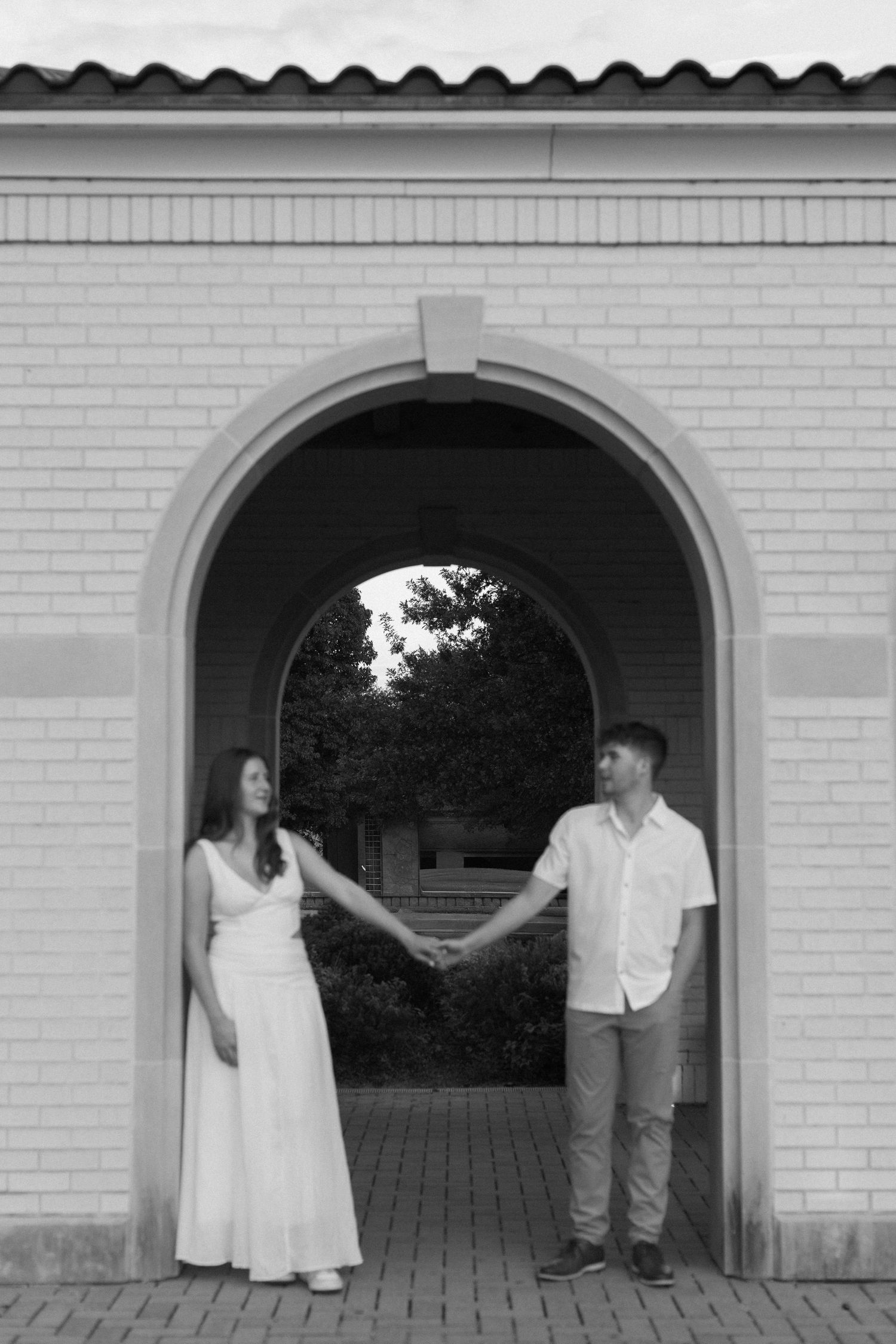 A couple playfully poses through arched openings in a white brick wall.