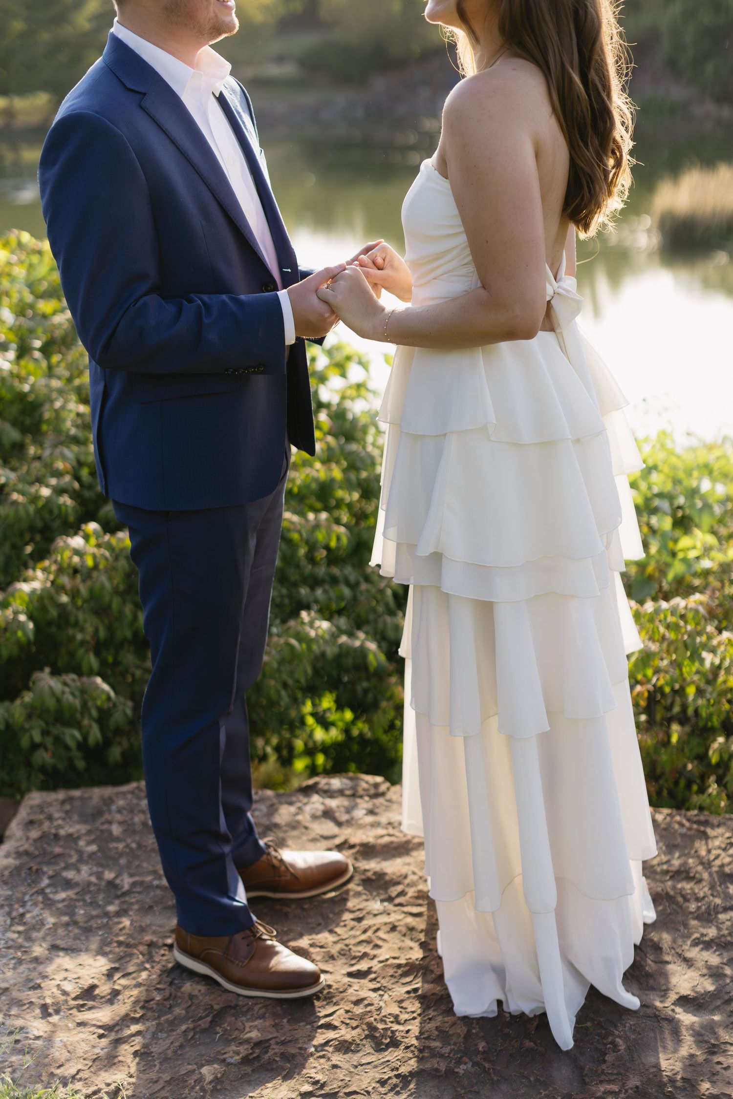 A couple in wedding attire stands by a lake at sunset, holding hands in an intimate moment.