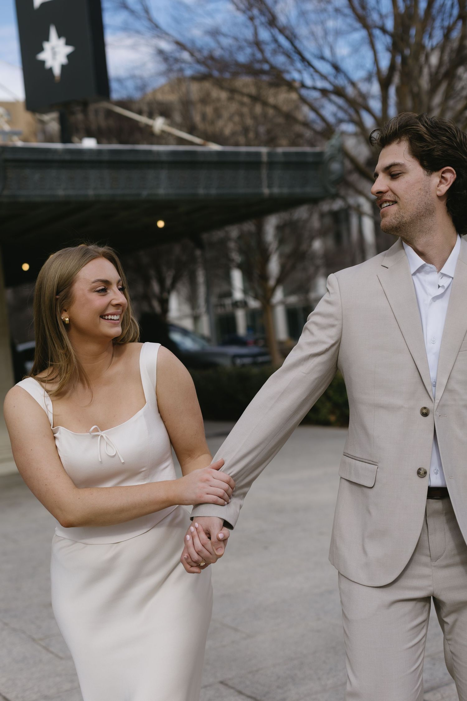 A couple in coordinating beige and white formal attire share a playful moment on a city sidewalk.