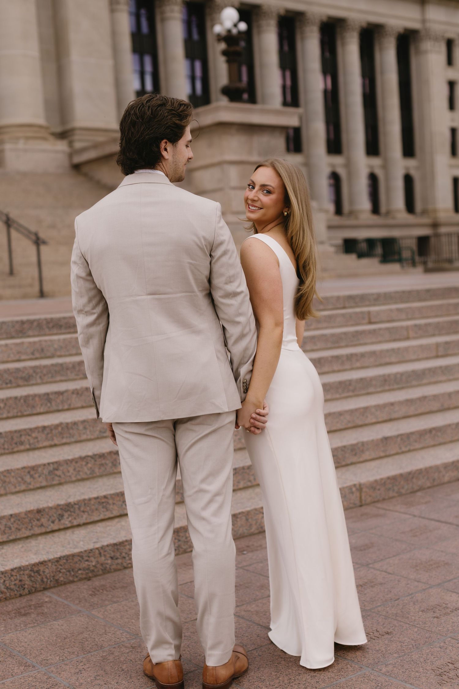 A couple in formal attire poses on grand stone steps, with the person in a white dress looking back at their partner.