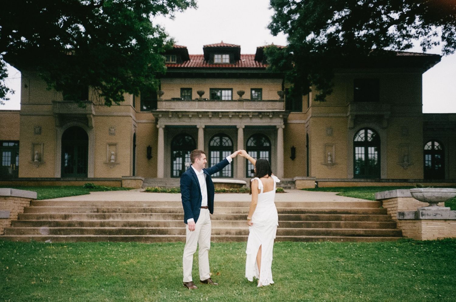 Couple in wedding attire standing on lawn in front of historic stone building.