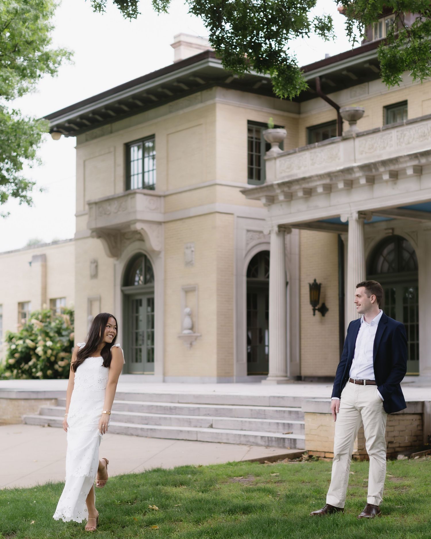 Couple poses playfully outside grand stone building with columns and arched architecture.