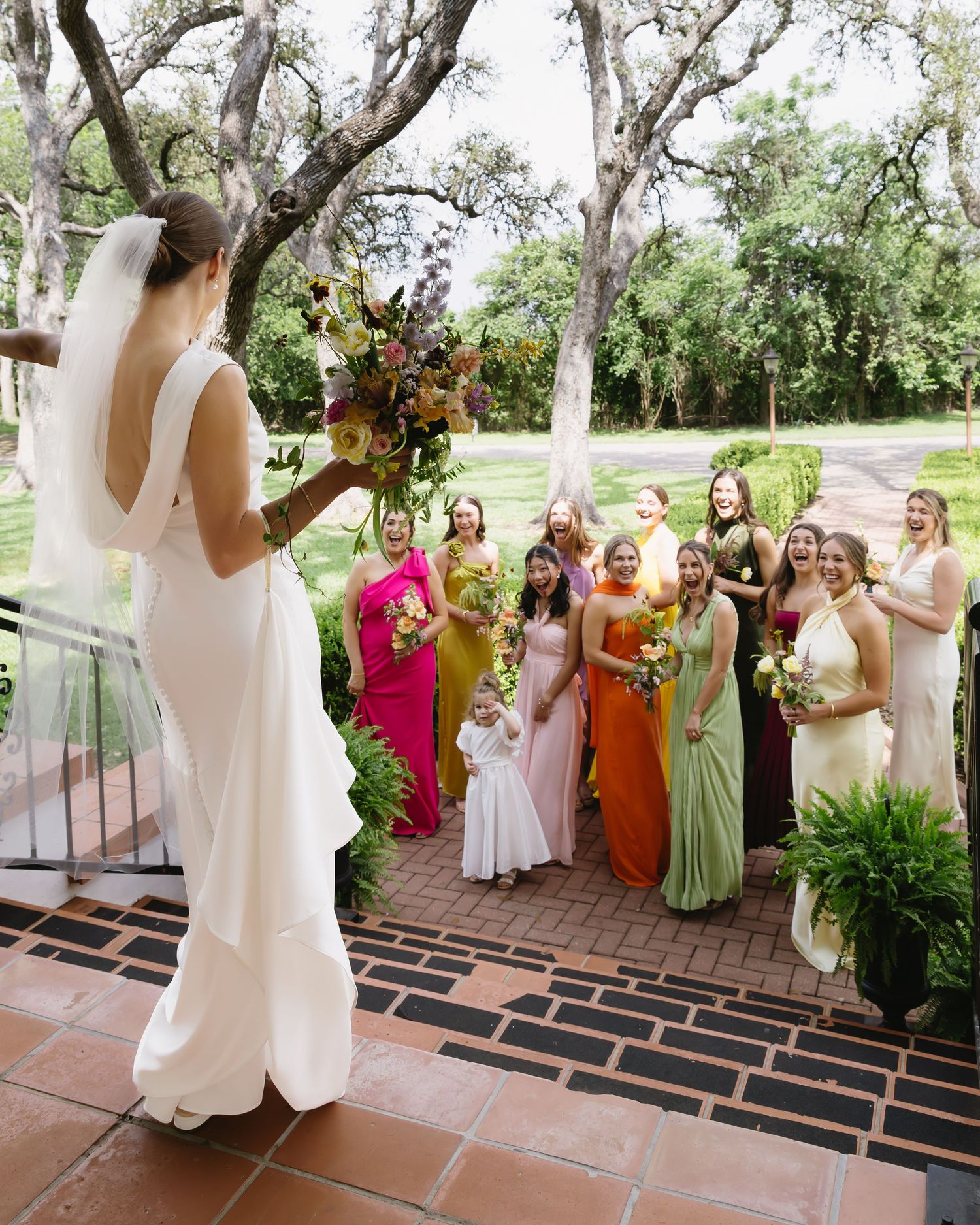 A wedding celebration with attendees in colorful dresses gathering on a brick patio surrounded by trees.