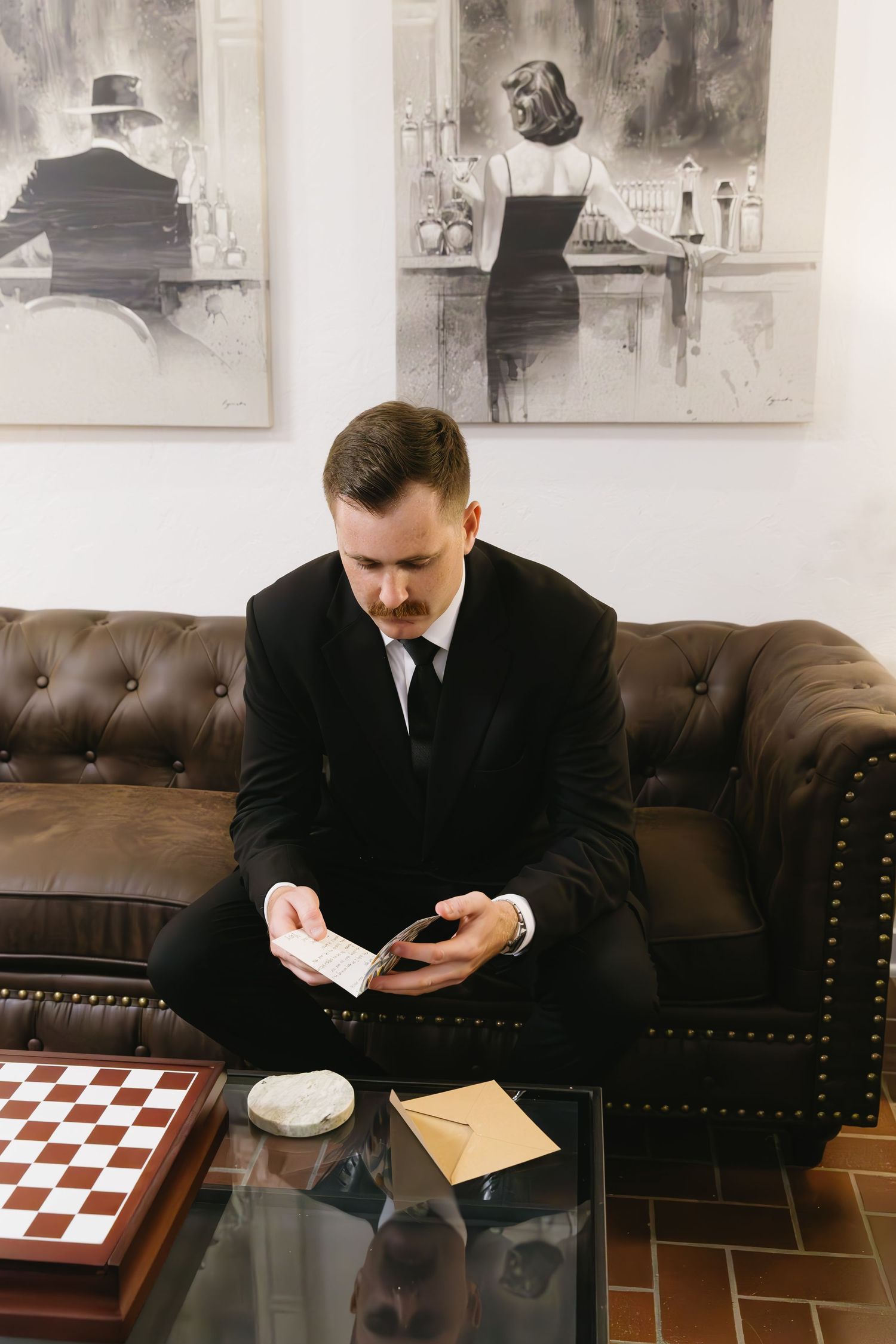 Formal gentleman sitting on leather couch checking phone near vintage artwork.