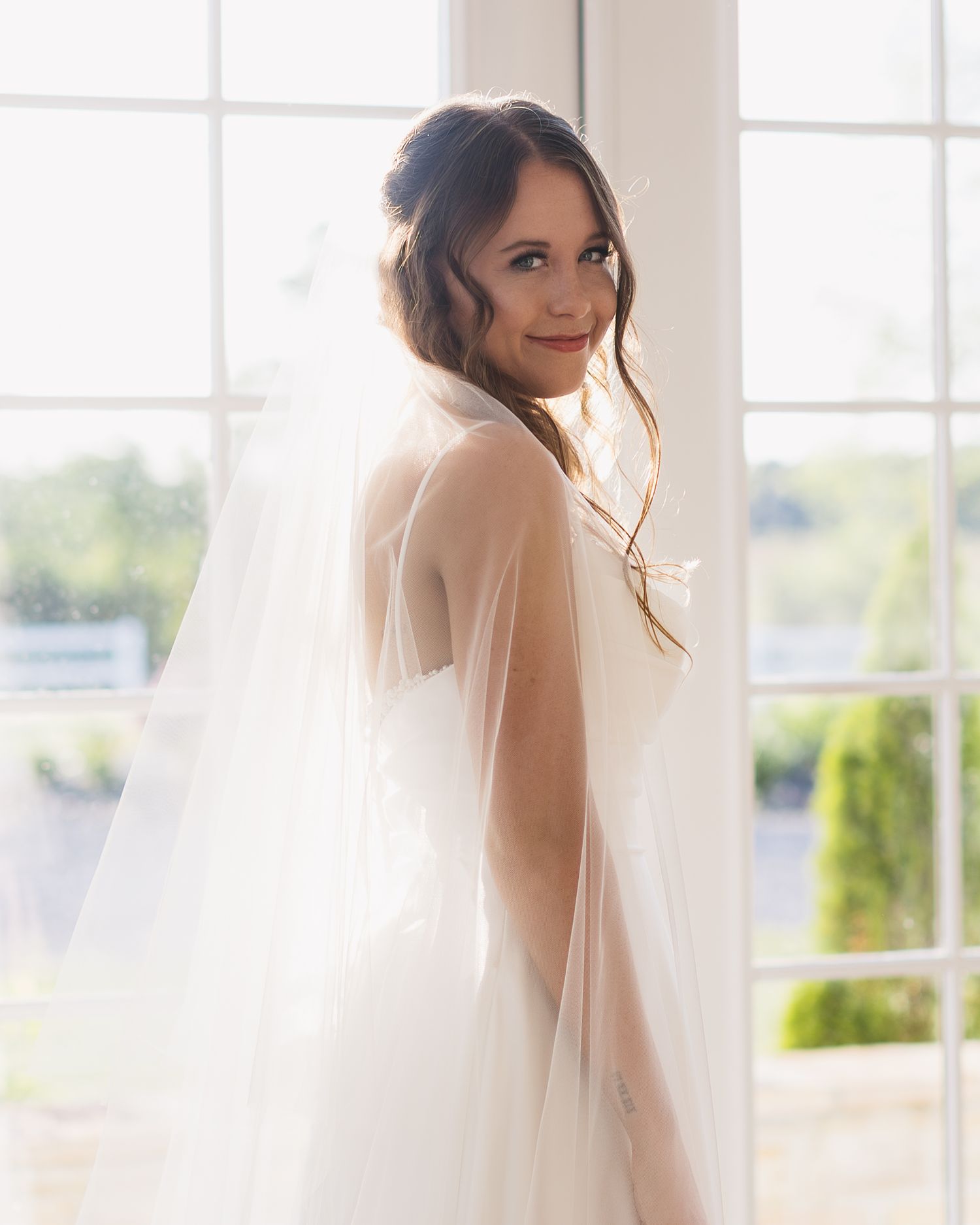 A bride in a flowing white dress poses near a window with natural light creating an ethereal effect.