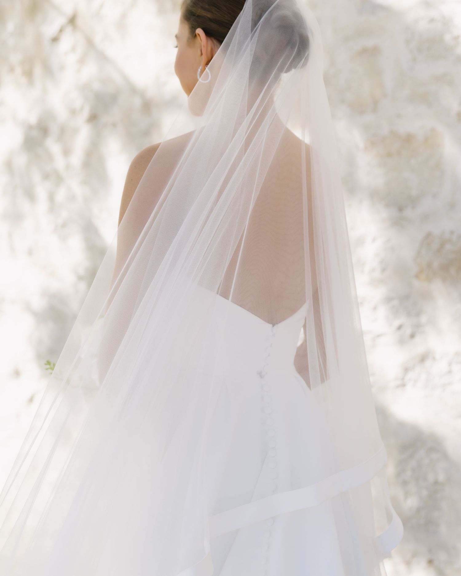 Back view of bride in strapless wedding dress with flowing veil against soft light background.