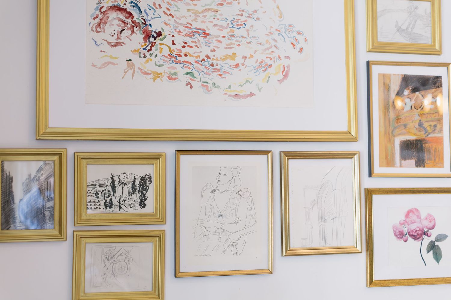 A gallery wall features multiple framed artworks and sketches in gold frames arranged on a white wall.