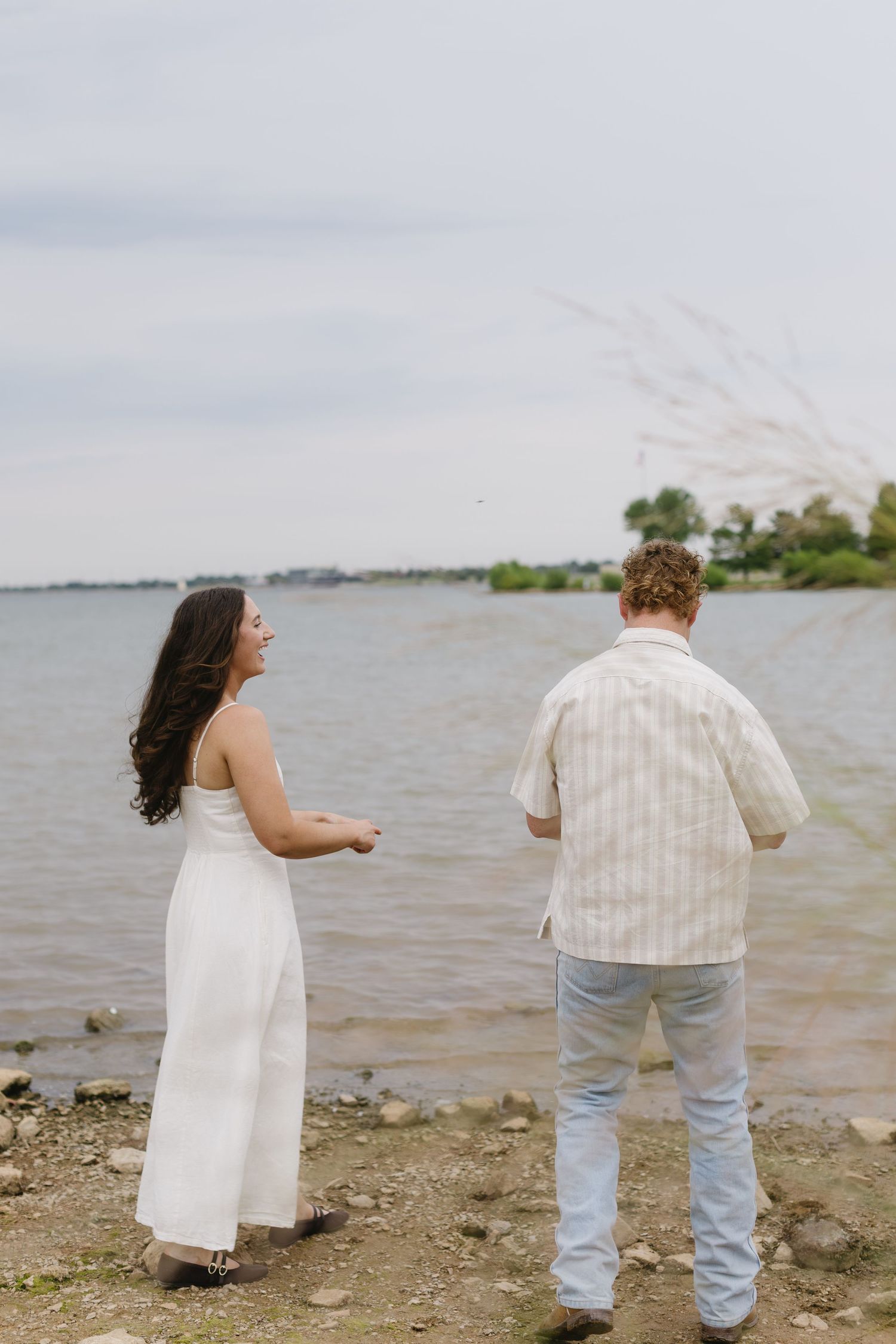 A couple stands by the water's edge on a rocky shoreline wearing casual attire on an overcast day.