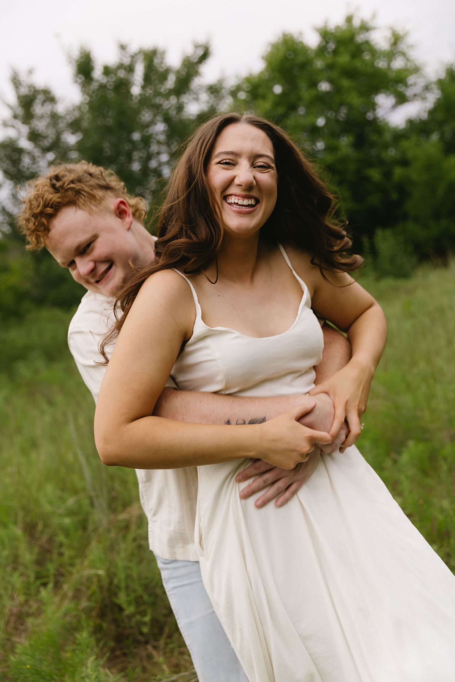 Two people share laughter and embrace while standing in a summer meadow with green trees.