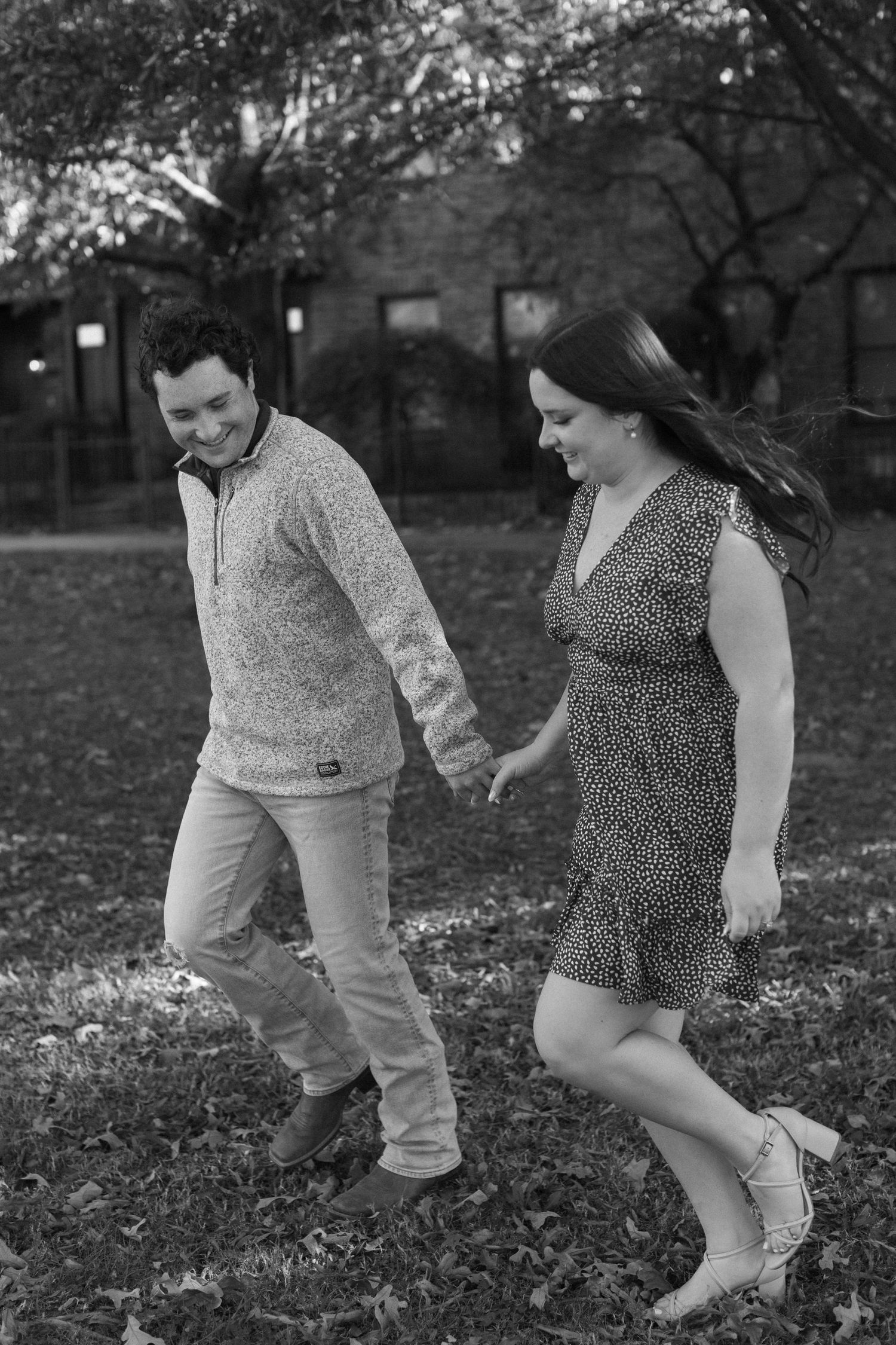 Two people playfully walk through fallen leaves in a park during an autumn photo session.