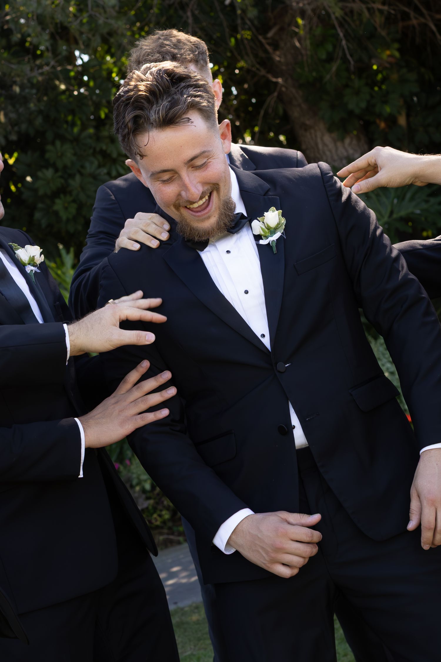 Groomsmen in black tuxedos share playful moments and laughter during wedding photo session.