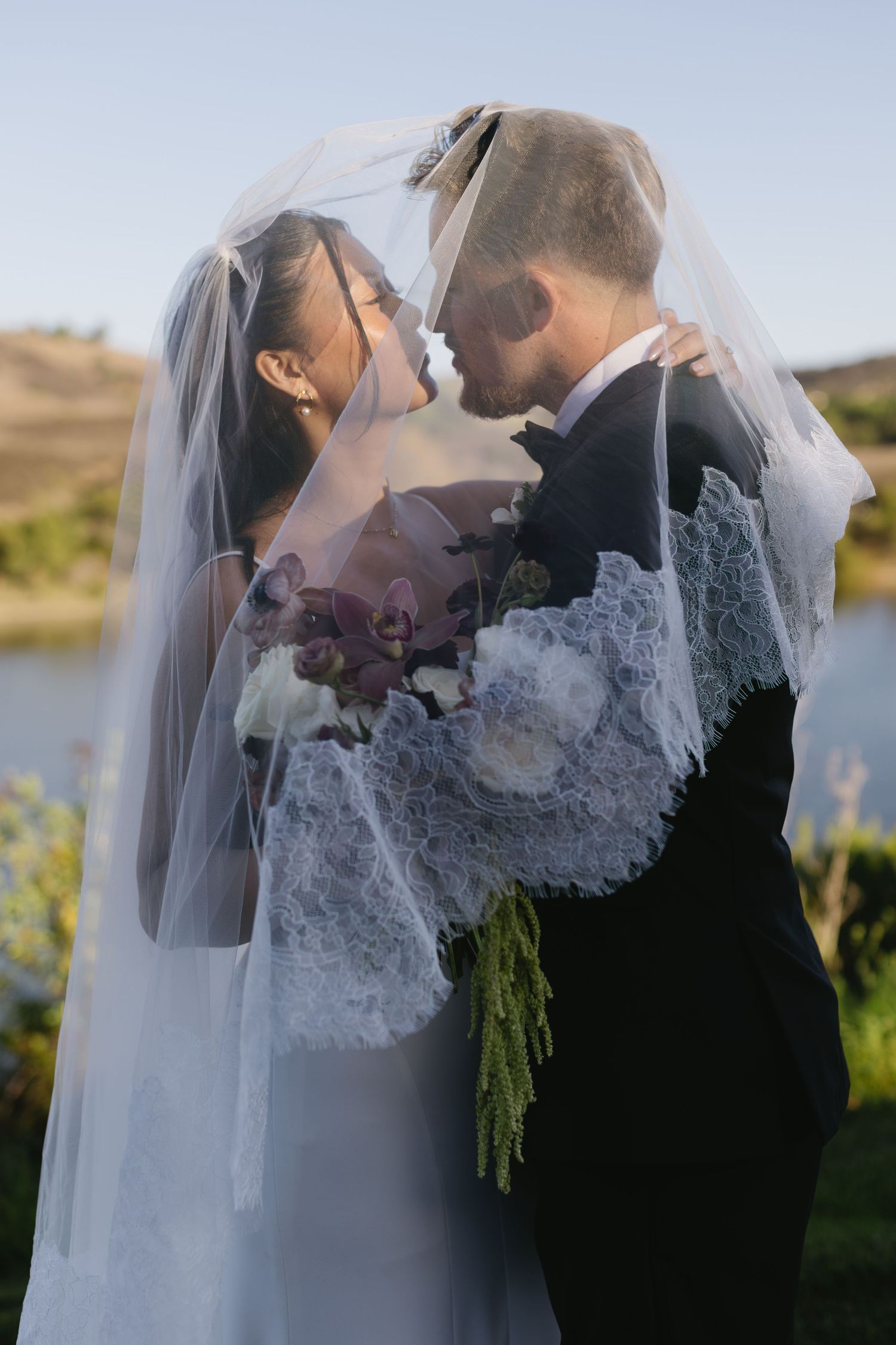 A romantic wedding moment captured by a lake with delicate lace veil details.