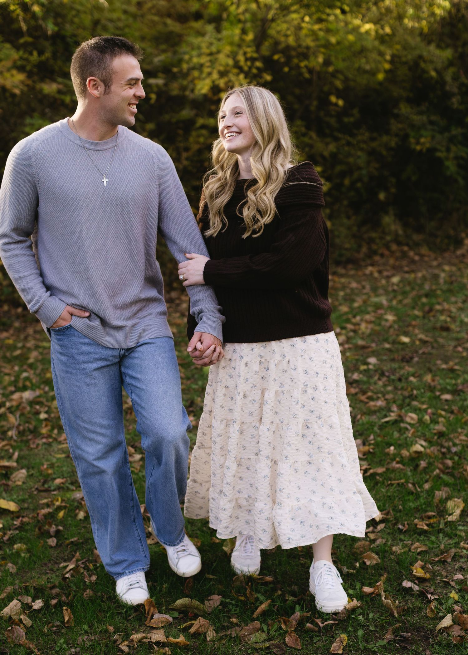 A couple shares a romantic moment while walking through fallen autumn leaves in a park setting.