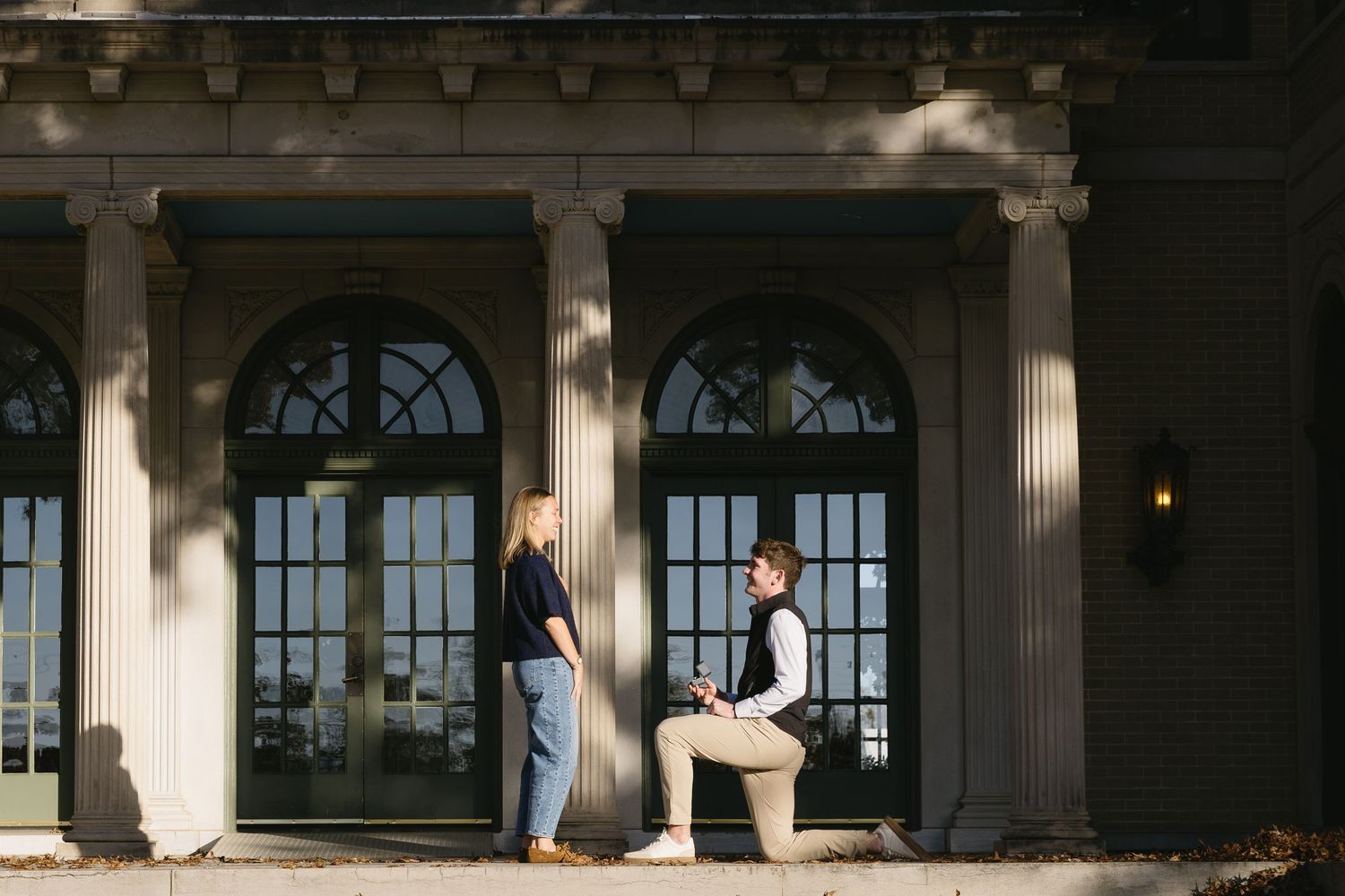 A romantic proposal moment unfolds on a grand porch with elegant columns and arched windows in the background.