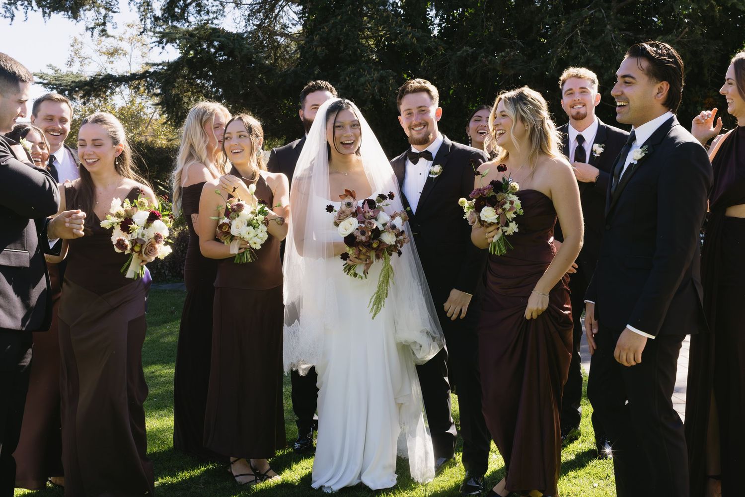 Wedding party members in brown suits and white dresses gather outdoors for formal photos on a sunny day.
