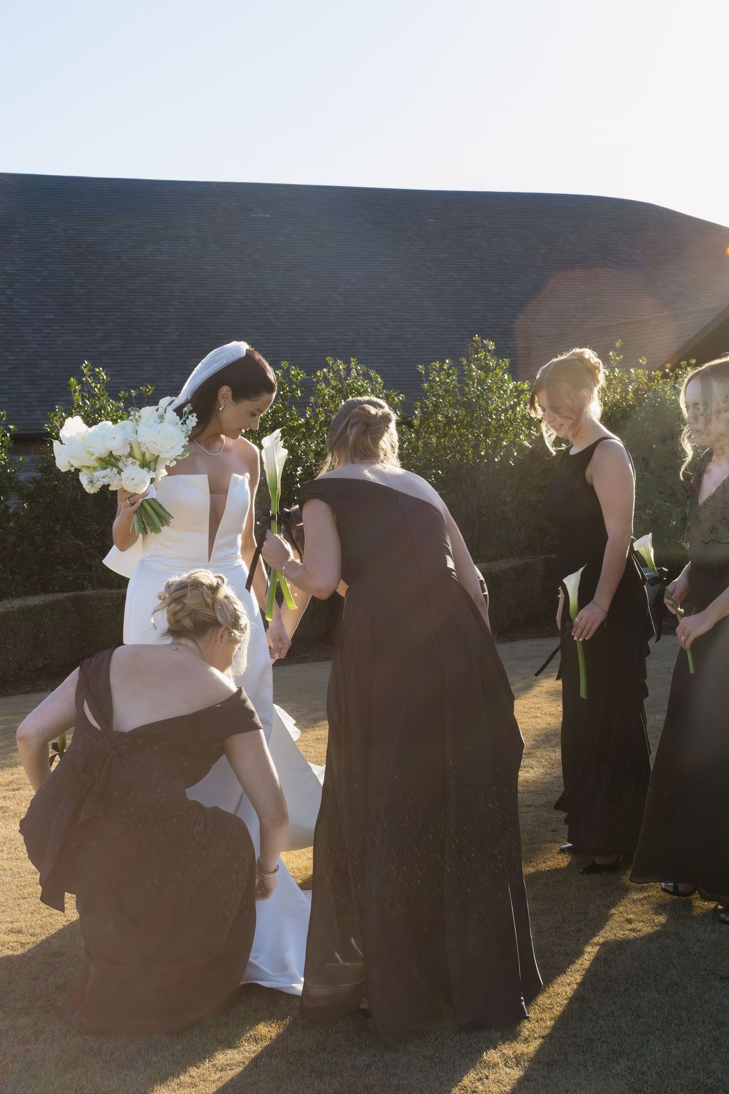 Wedding party gathers outdoors in formal attire with black dresses and white bouquet against hillside backdrop.