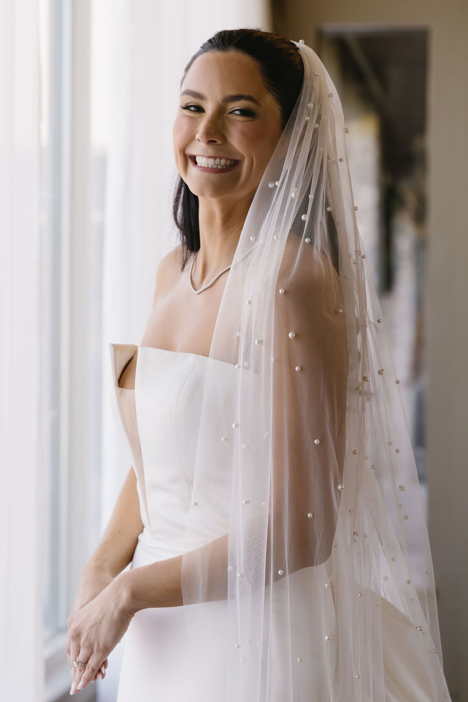 A bride in a white wedding dress and pearl-studded veil smiling by a window.