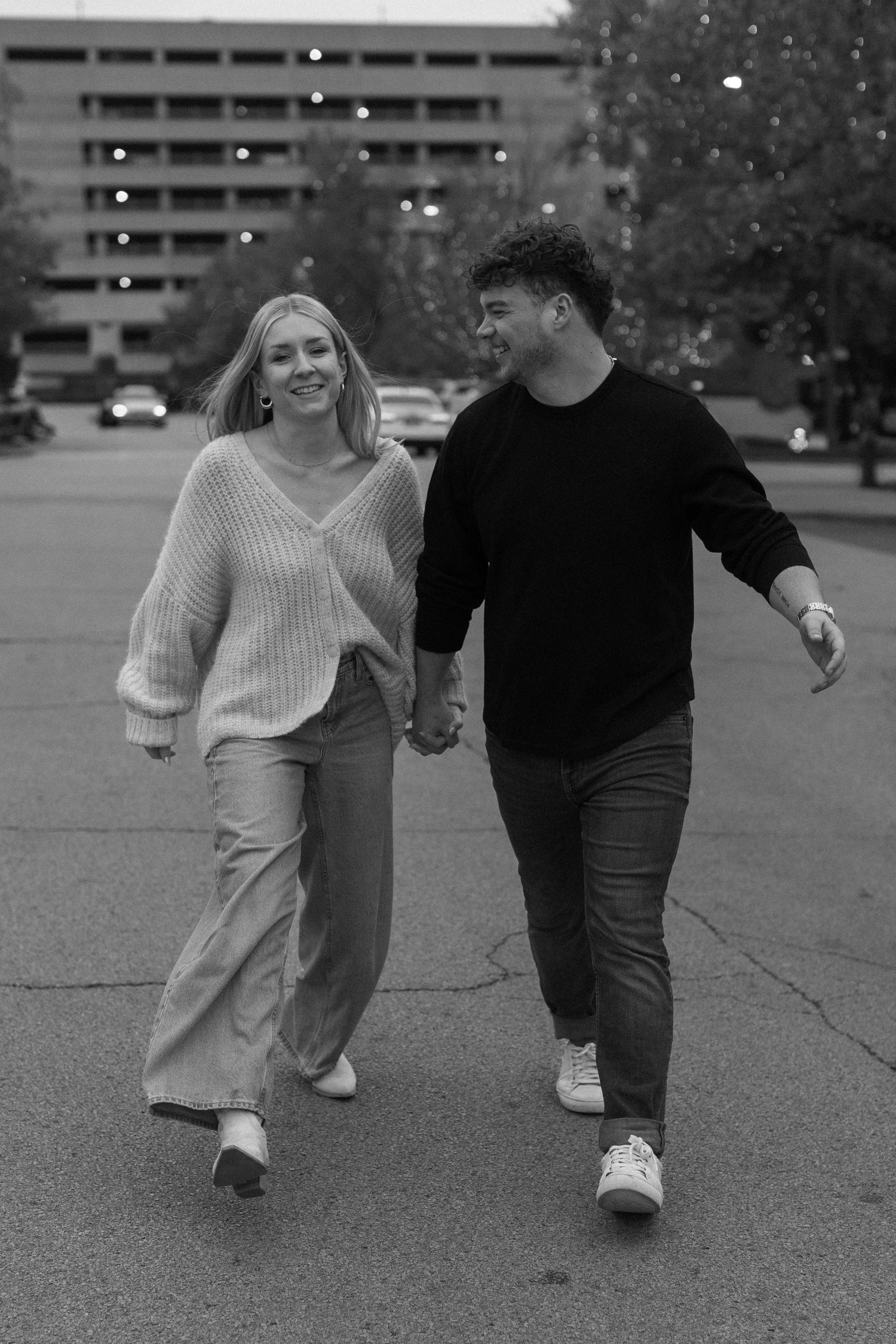 A couple walks playfully through a parking lot in black and white photography.