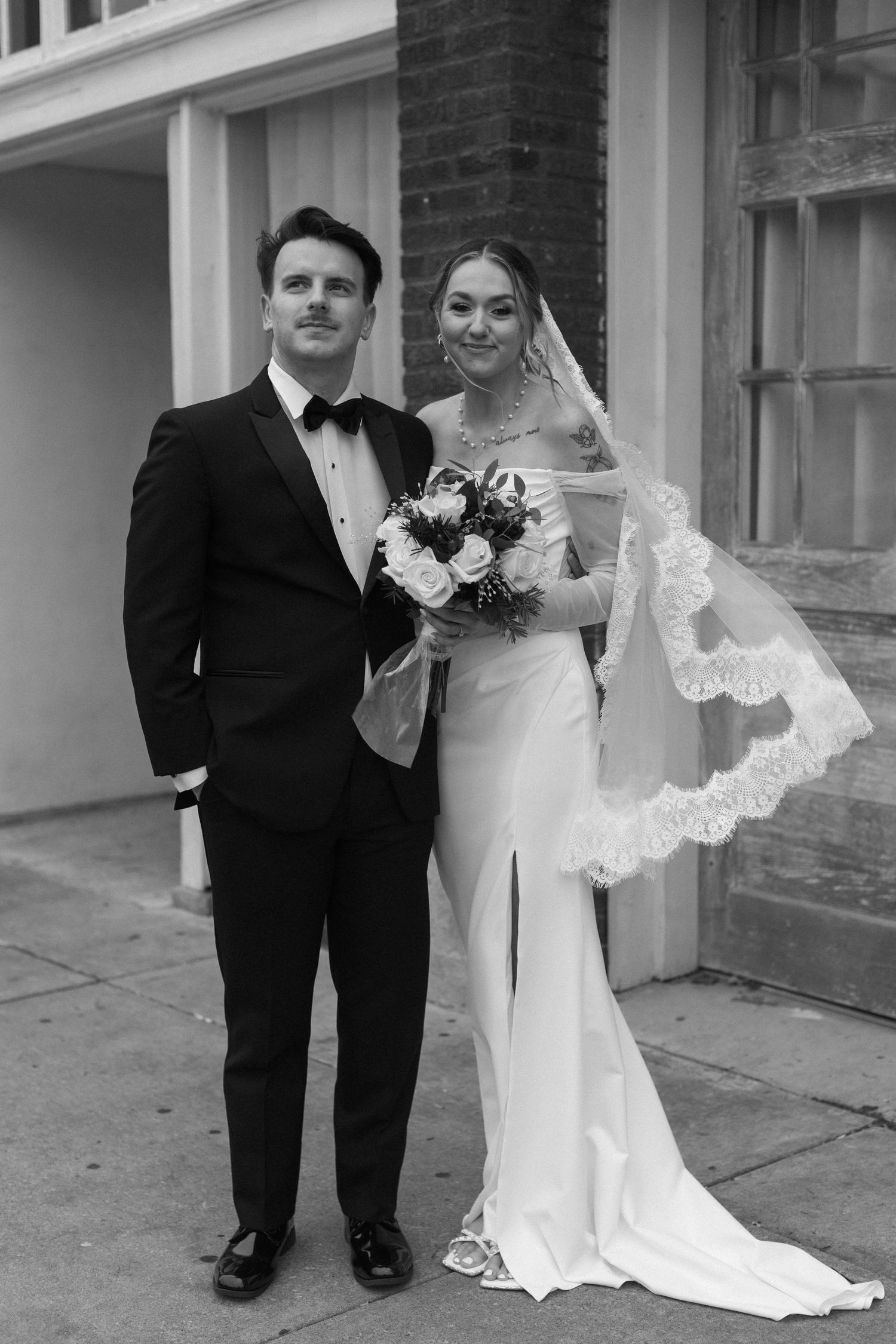 Black and white portrait of newlyweds standing together against a brick wall backdrop.