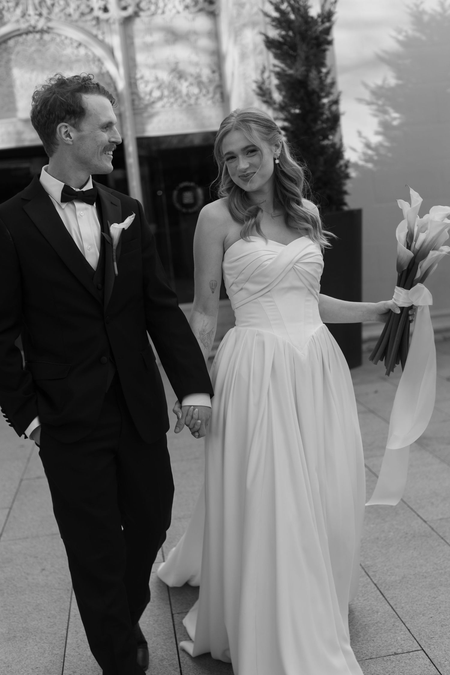 A bride in a flowing white gown and groom in black tuxedo share candid moments outdoors.