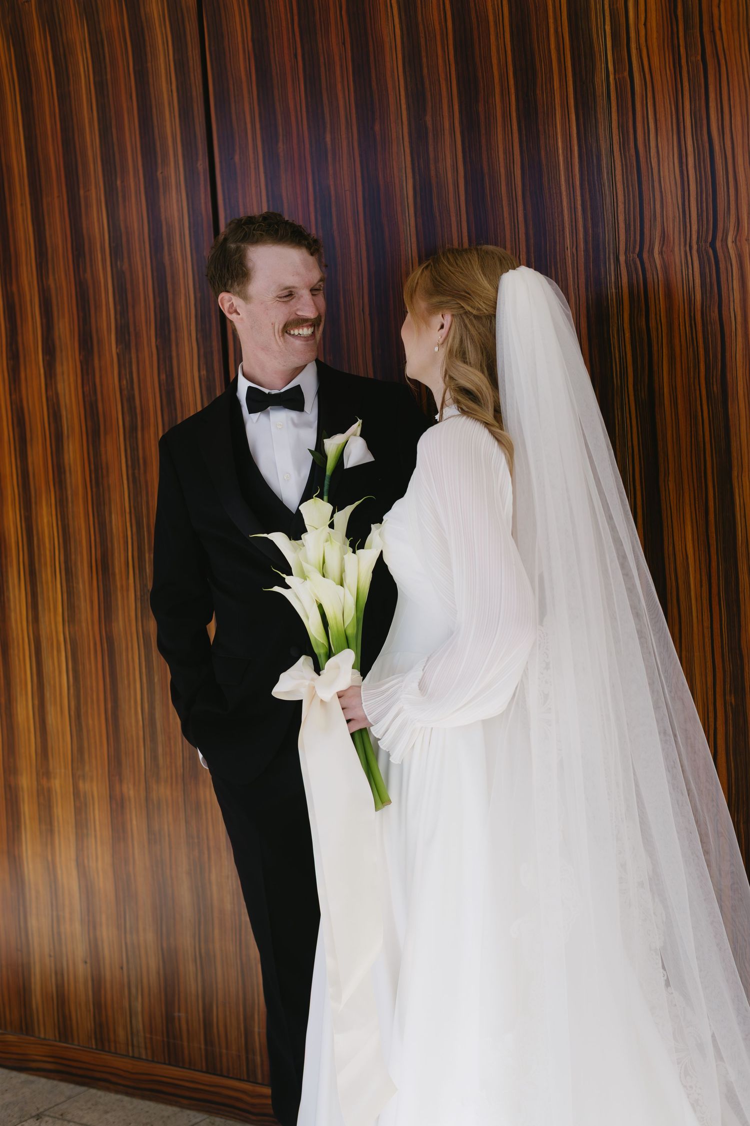 Wedding couple sharing intimate moments with white calla lily bouquet against wood paneled wall.