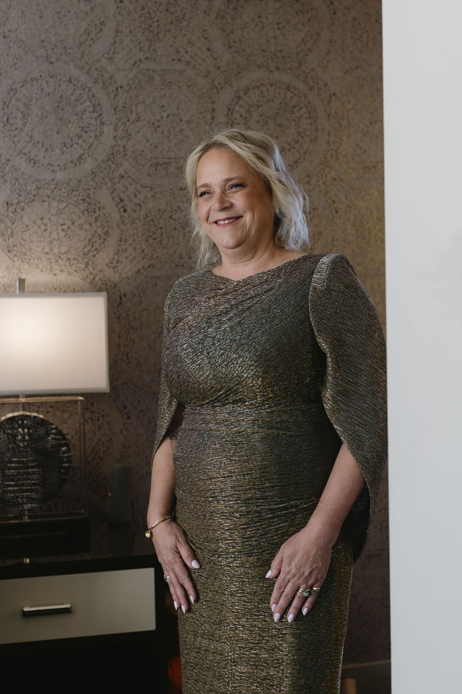 Woman wearing a sparkly green evening dress poses against a textured wall with elegant lighting.