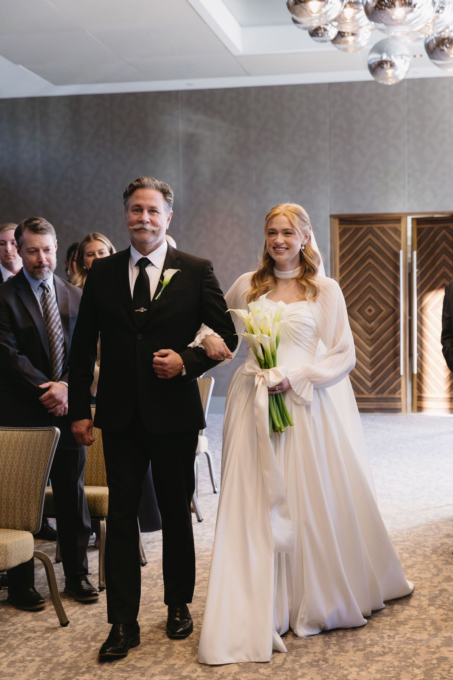 Wedding processional in elegant indoor venue with crystal chandeliers overhead.