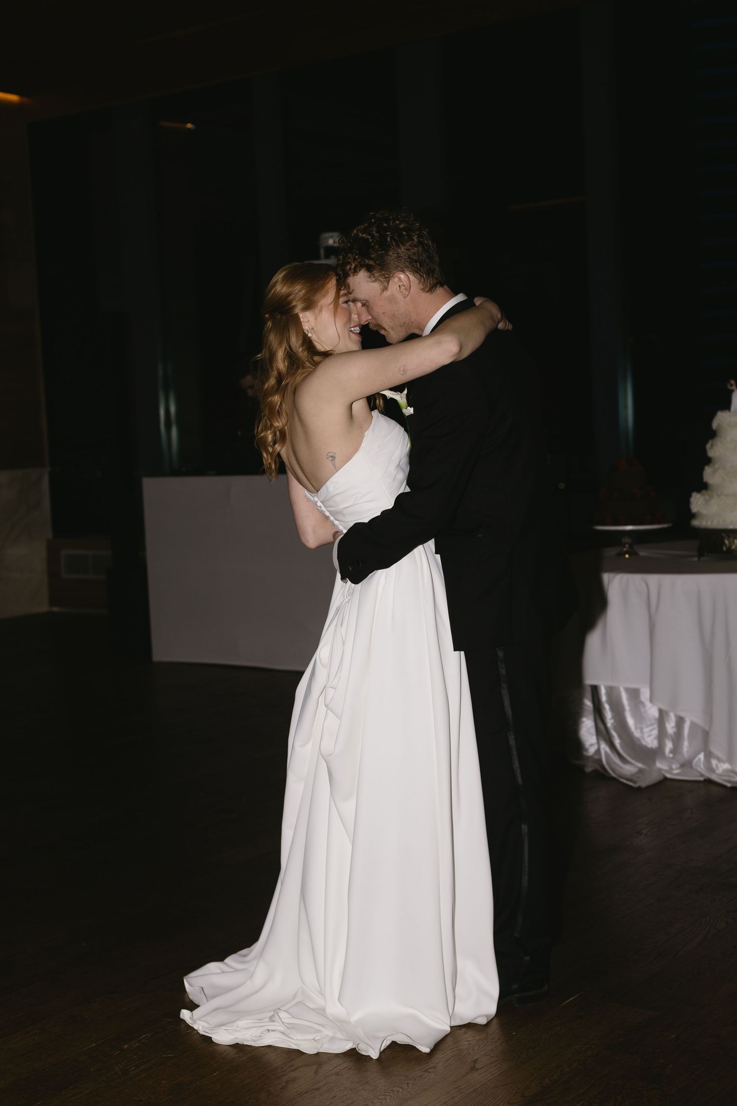 A couple shares an intimate dance in a dimly lit reception hall with white tablecloths.