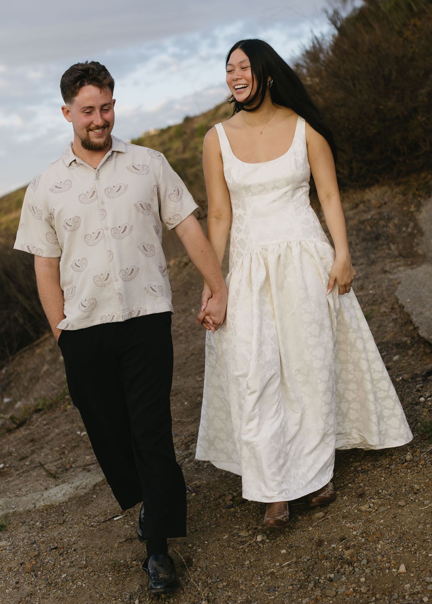 A couple poses together on a hillside trail wearing coordinating white and beige outfits at sunset.