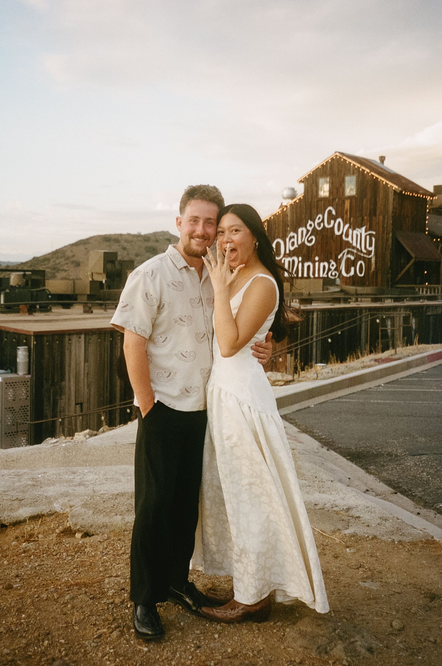 A couple shares romantic moments in front of rustic wooden buildings at sunset in a desert ghost town setting.
