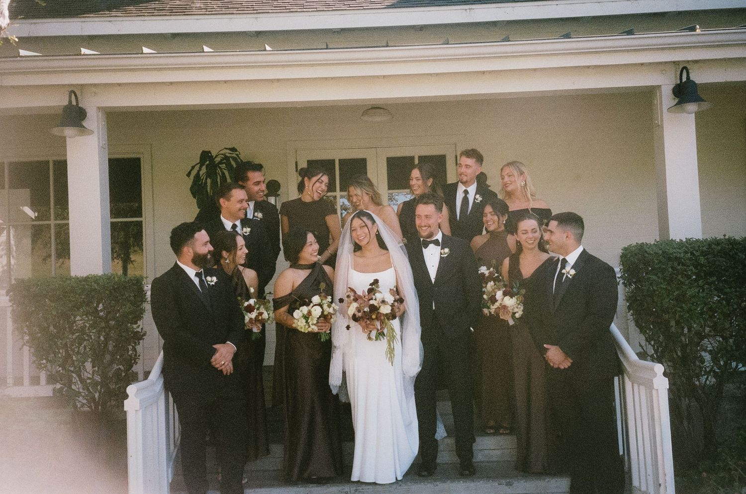 Wedding party in formal attire poses on a white columned porch with autumn floral bouquets.
