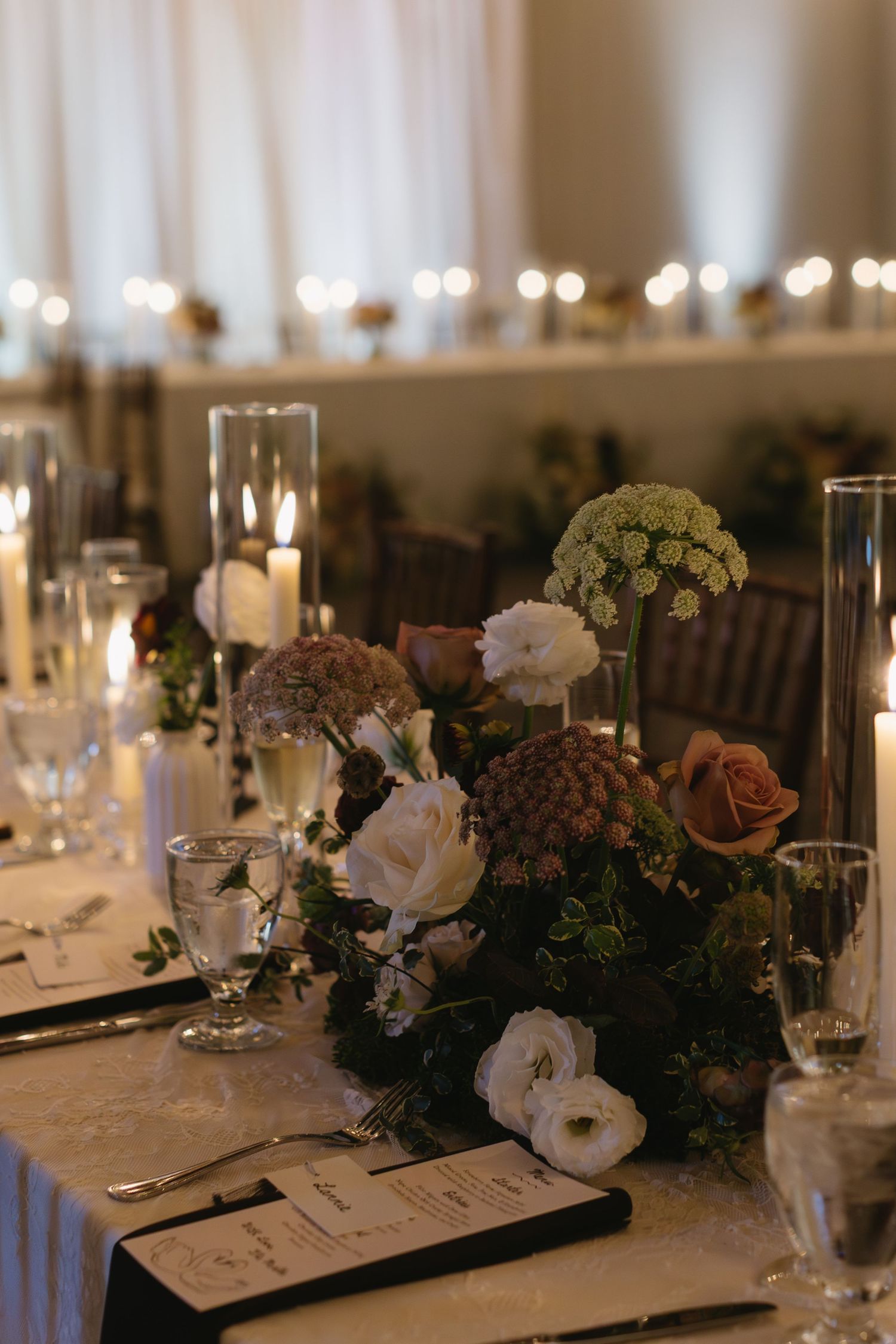Romantic reception table setting with floral centerpieces, candles, and glassware in warm lighting.